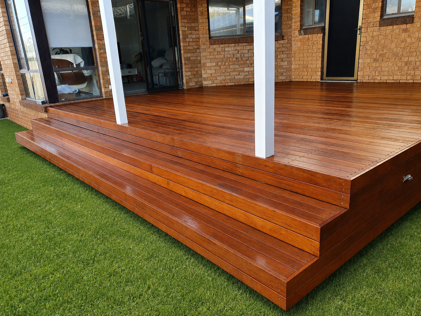 A wooden deck attached to a brick house, with stairs leading down to a lawn. The deck has a white post and a black door.