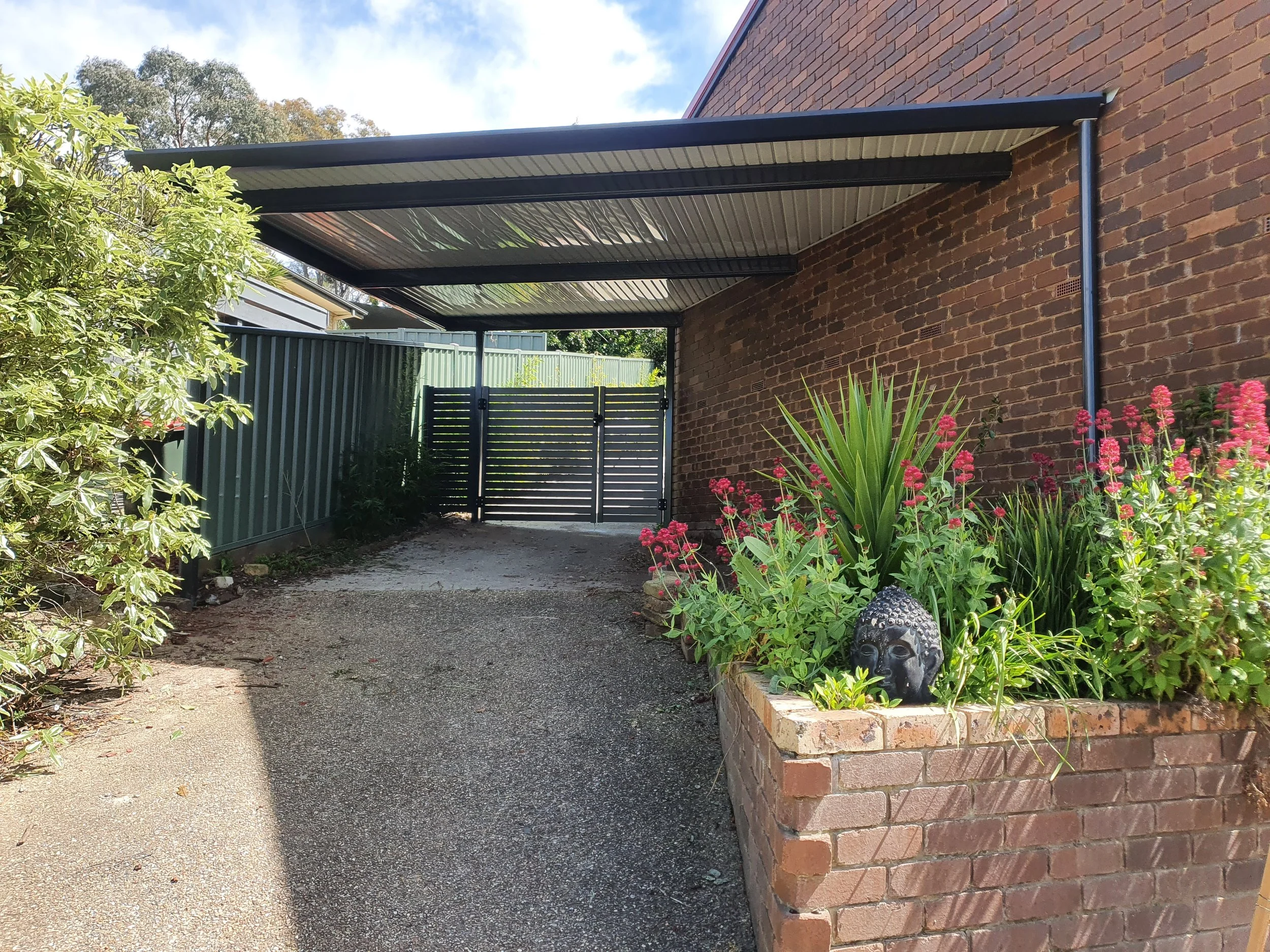 A backyard garden area with flowering plants, a brick wall, a metal fence, and a metal and plastic roof over a walkway.