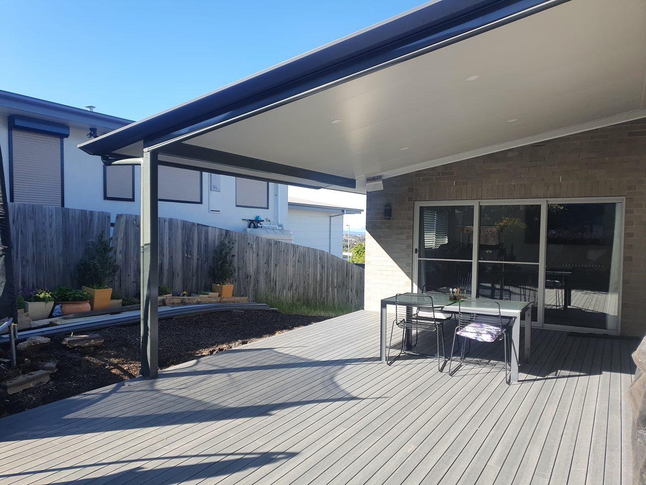 A backyard patio with a table and chairs, attached to a house with large sliding glass doors, and a covered roof with recessed lighting. A wooden fence and some plant pots are visible in the background.