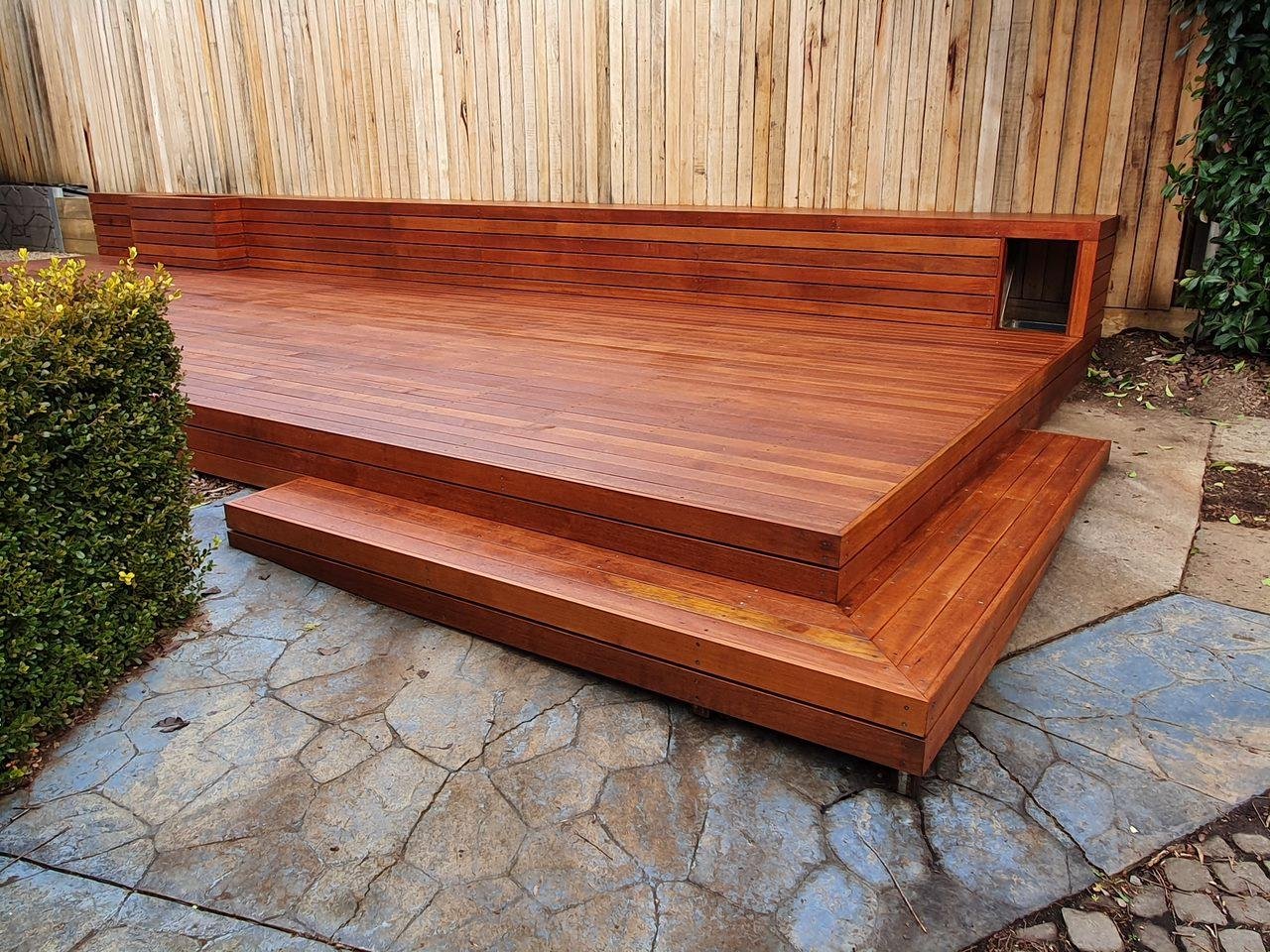 Outdoor wooden deck with built-in seating, surrounded by stone paving, a bush on the left, and a wooden fence and greenery on the right.