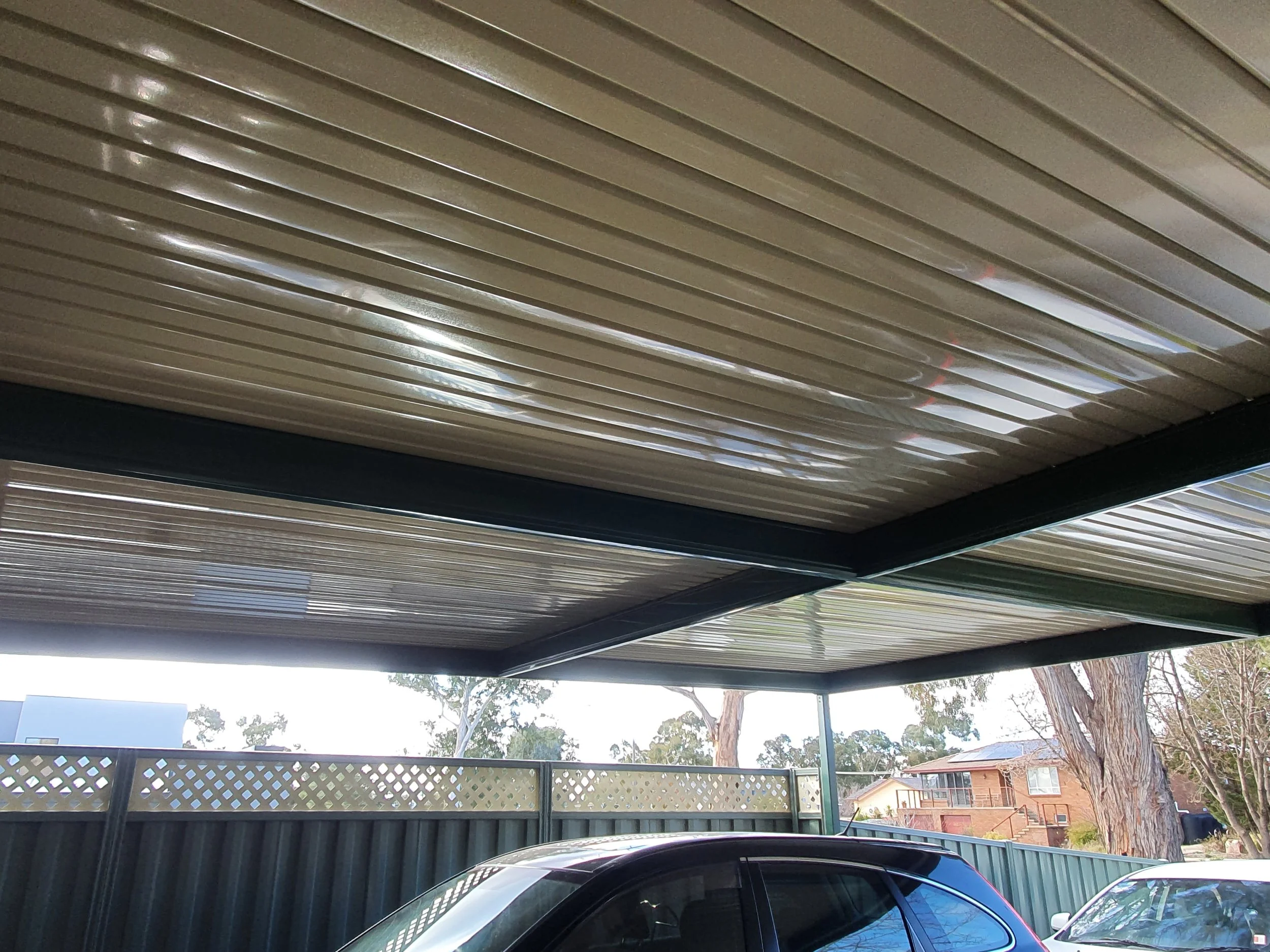 View of a carport with a metal slatted roof, part of a parked black car visible in the foreground, a green wooden fence, trees, neighboring houses, and a partly cloudy sky.