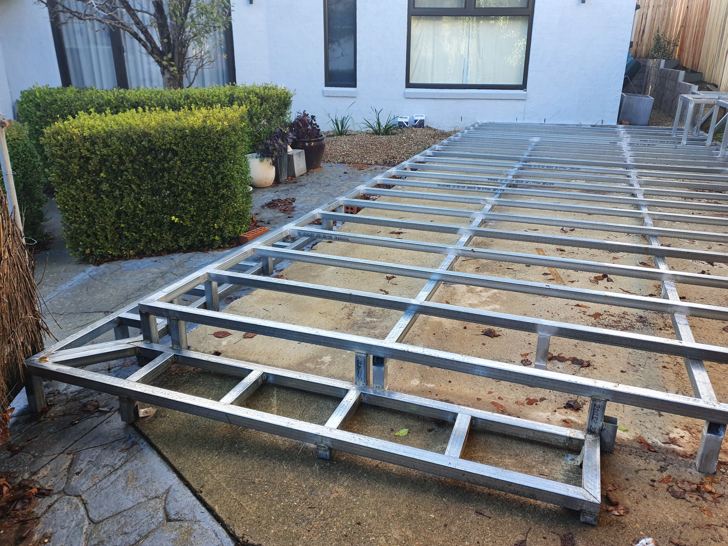 Metal framework for a deck in a backyard, with bushes and a house in the background.
