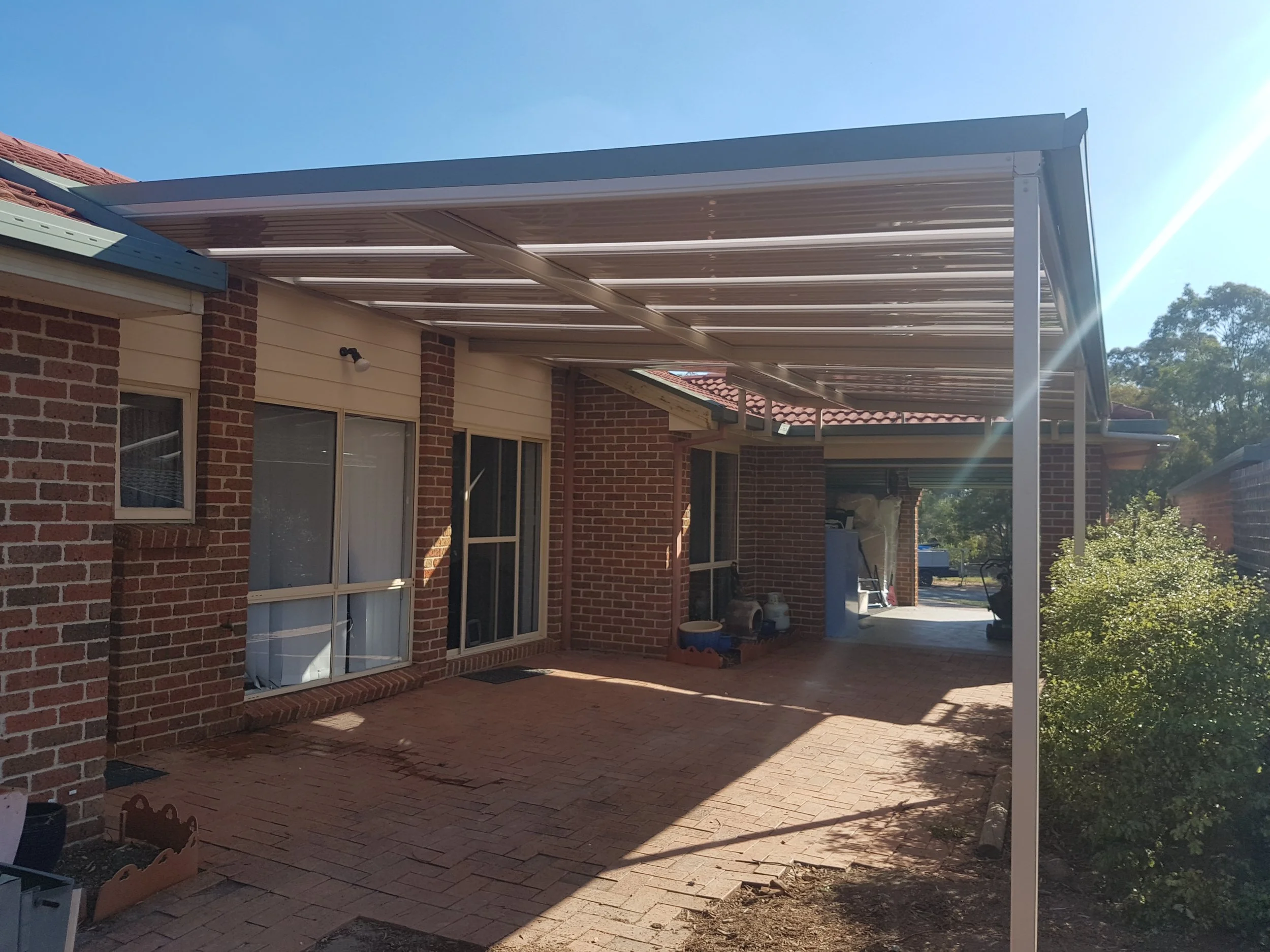 Backyard patio area with brick paving, brick and siding house wall, windows, and a covered shelter with a metal roof and support beams, surrounded by trees and plants under a clear blue sky.