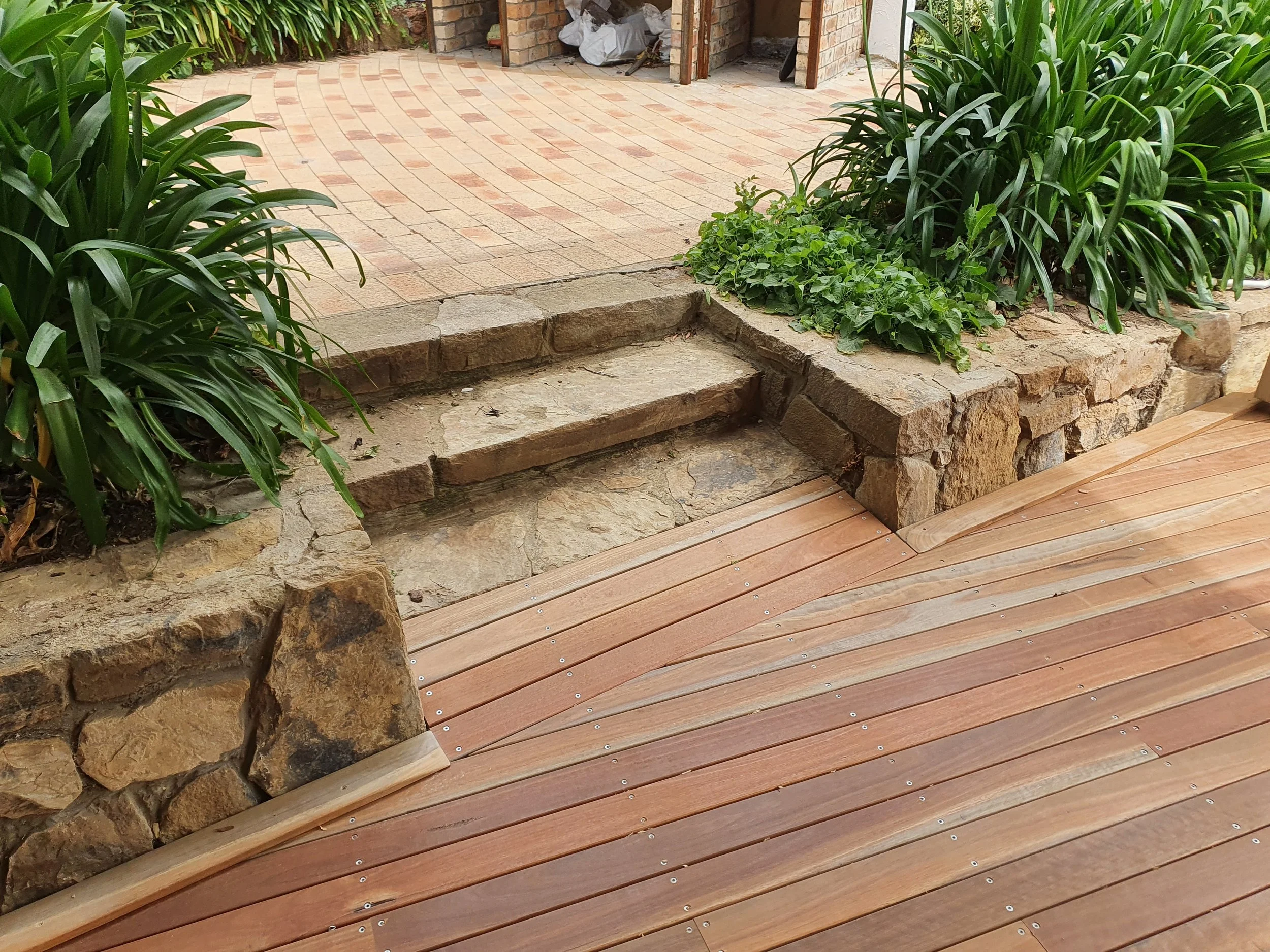 A stone step leading to a brick patio, with green plants on either side and an unfinished brick structure in the background.