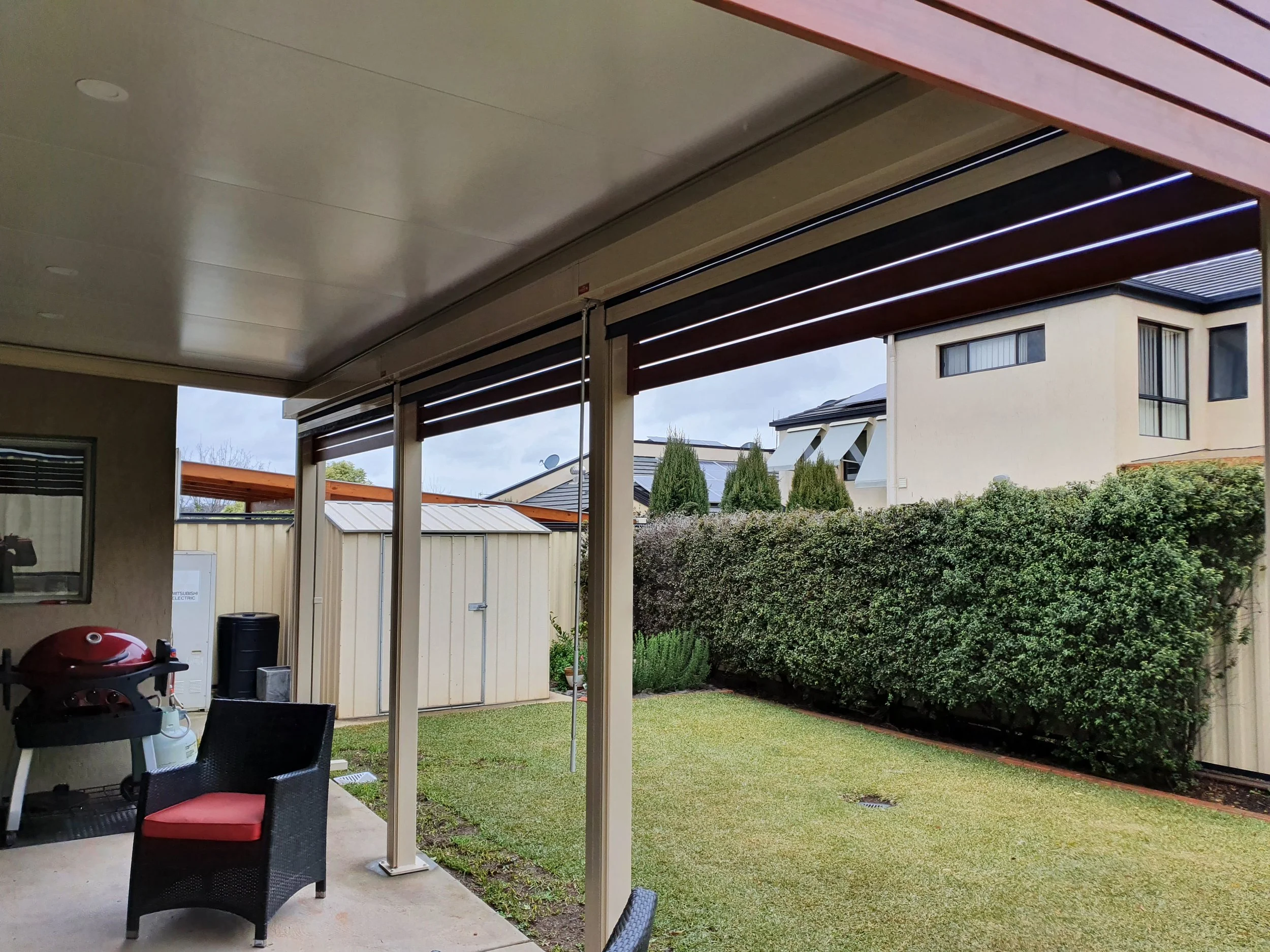 View of a backyard patio area with a covered roof, seating chairs, a grassy lawn, tall bushes, neighboring houses, and a cloudy sky.