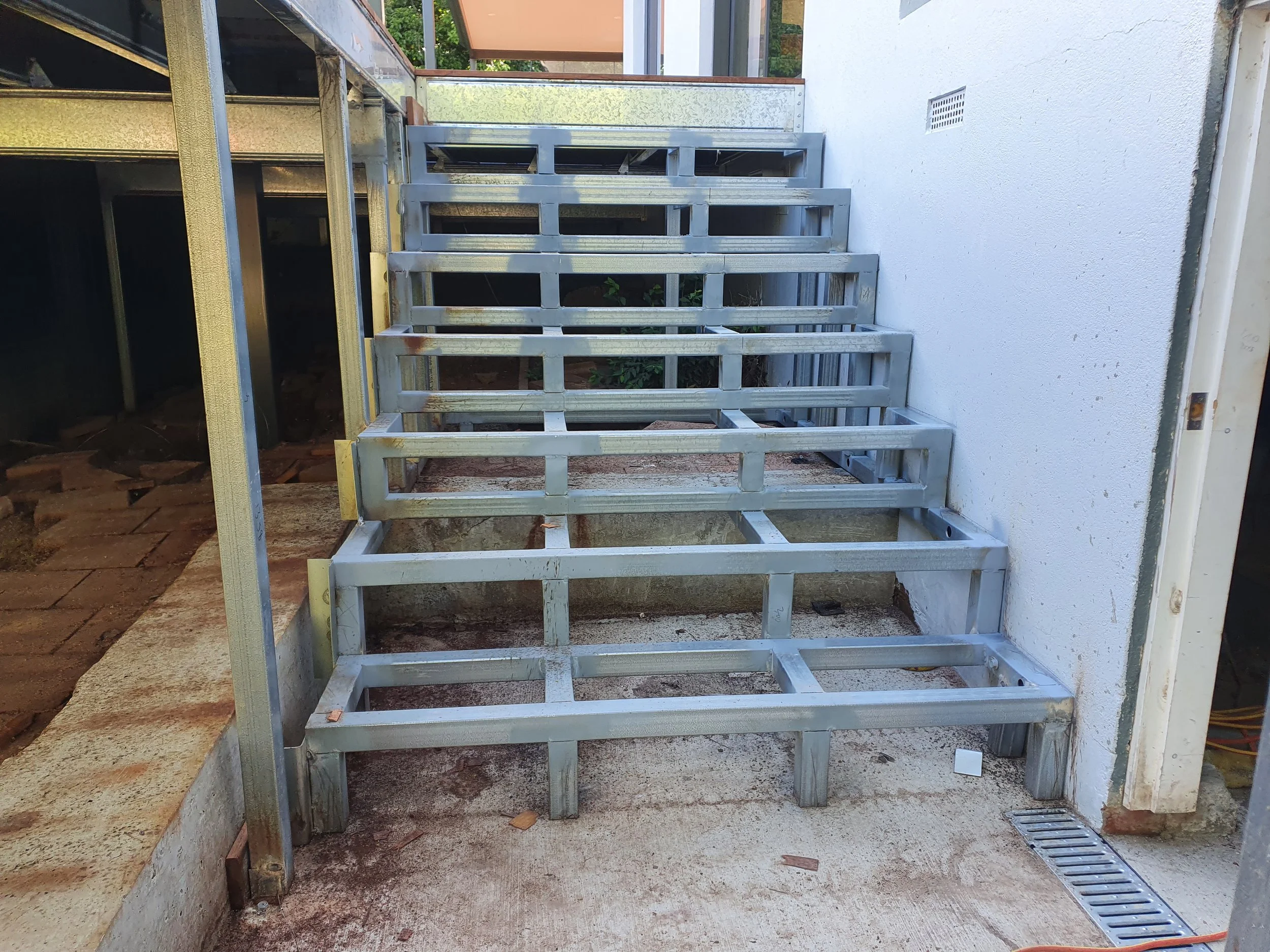 Steel framing for a stairway or balcony under construction adjacent to a white wall, with visible drainage grate on the ground.