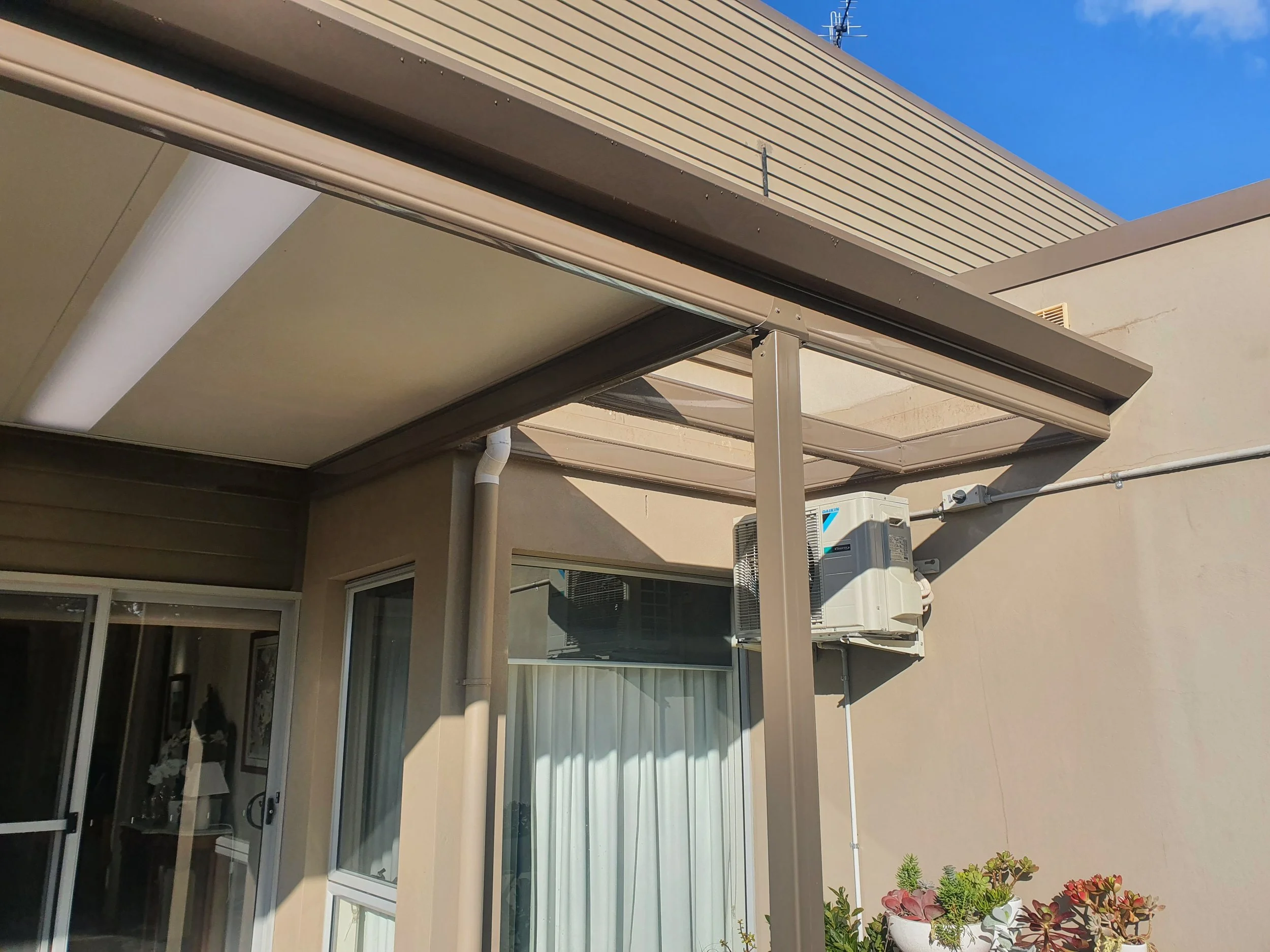A view of a house exterior showing a balcony with a beige or tan awning, sliding glass doors, and potted plants with succulents. The sky is clear and blue.