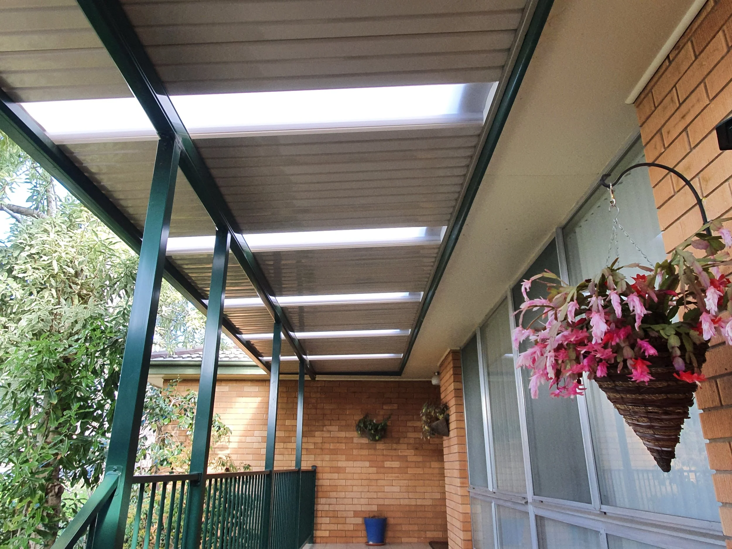 A covered outdoor patio area with green metal framing, a corrugated metal ceiling, brick walls, a large potted plant, and trees in the background.