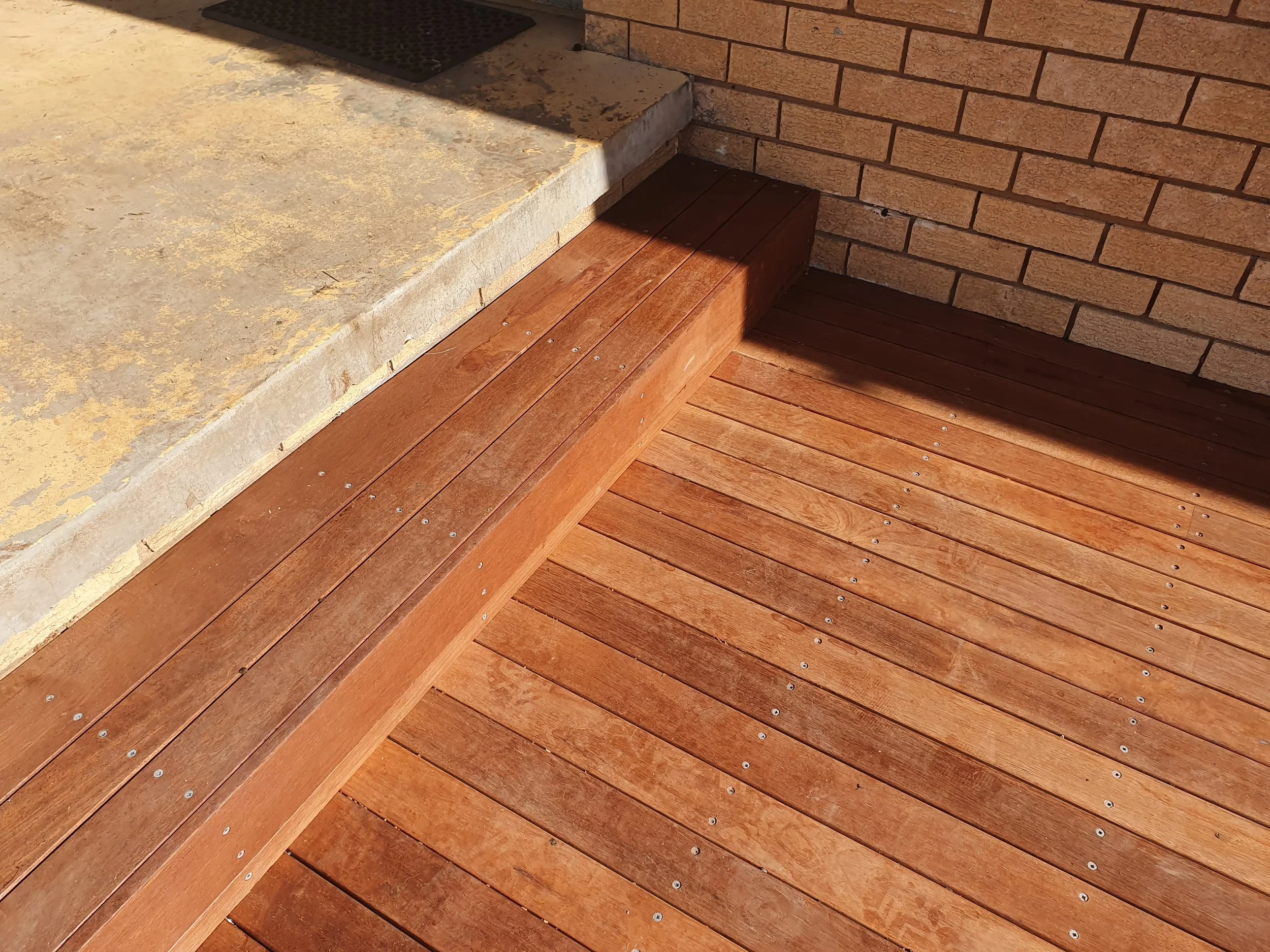 Close-up of a step area with beige concrete, a brick wall, and wooden flooring with metal screws, illuminated by sunlight.
