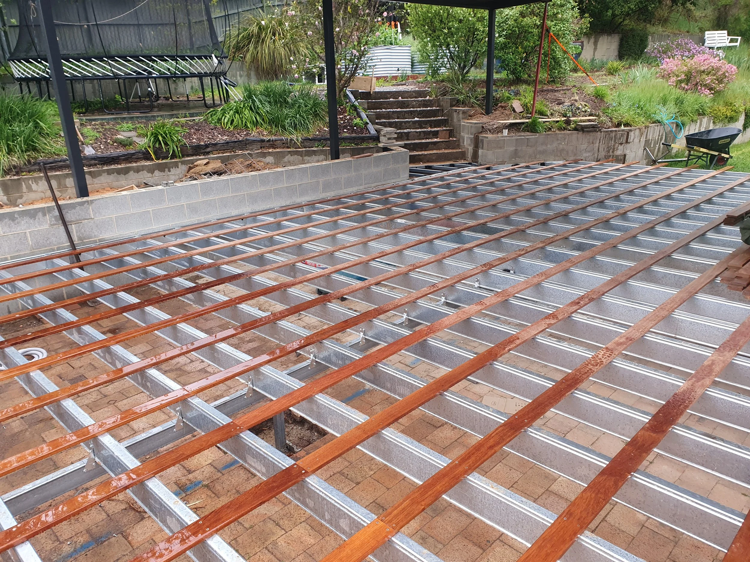 Construction site of a backyard deck with metal framing and wooden planks, garden stairs leading up to a landscaped area with plants, shrubs, and flowers.