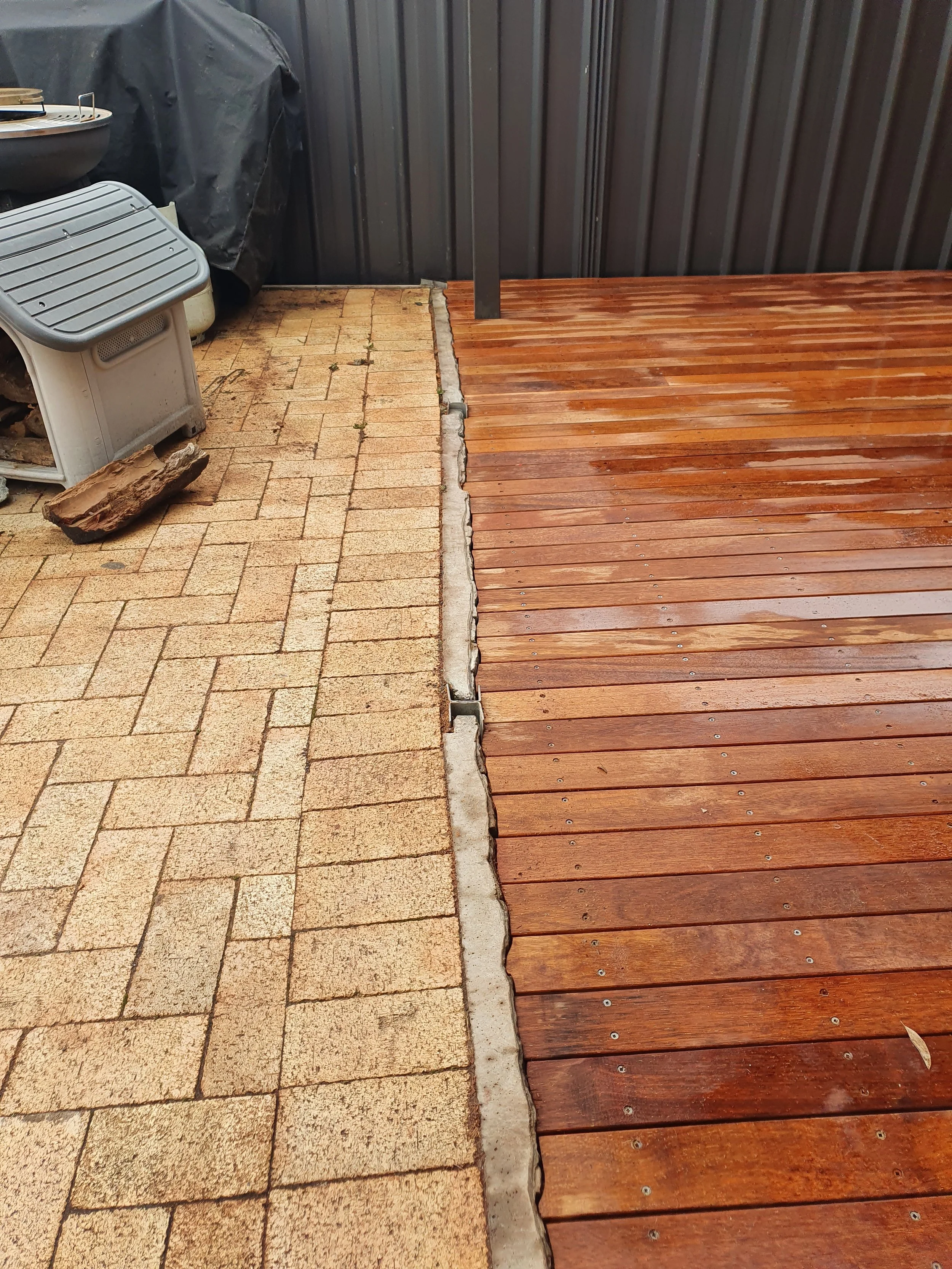 An outdoor area with a section of tan brick paving on the left and a wooden deck on the right, separated by a row of concrete pavers. In the background, there is a black covered grill, a plastic stool, and a large piece of wood on the ground.