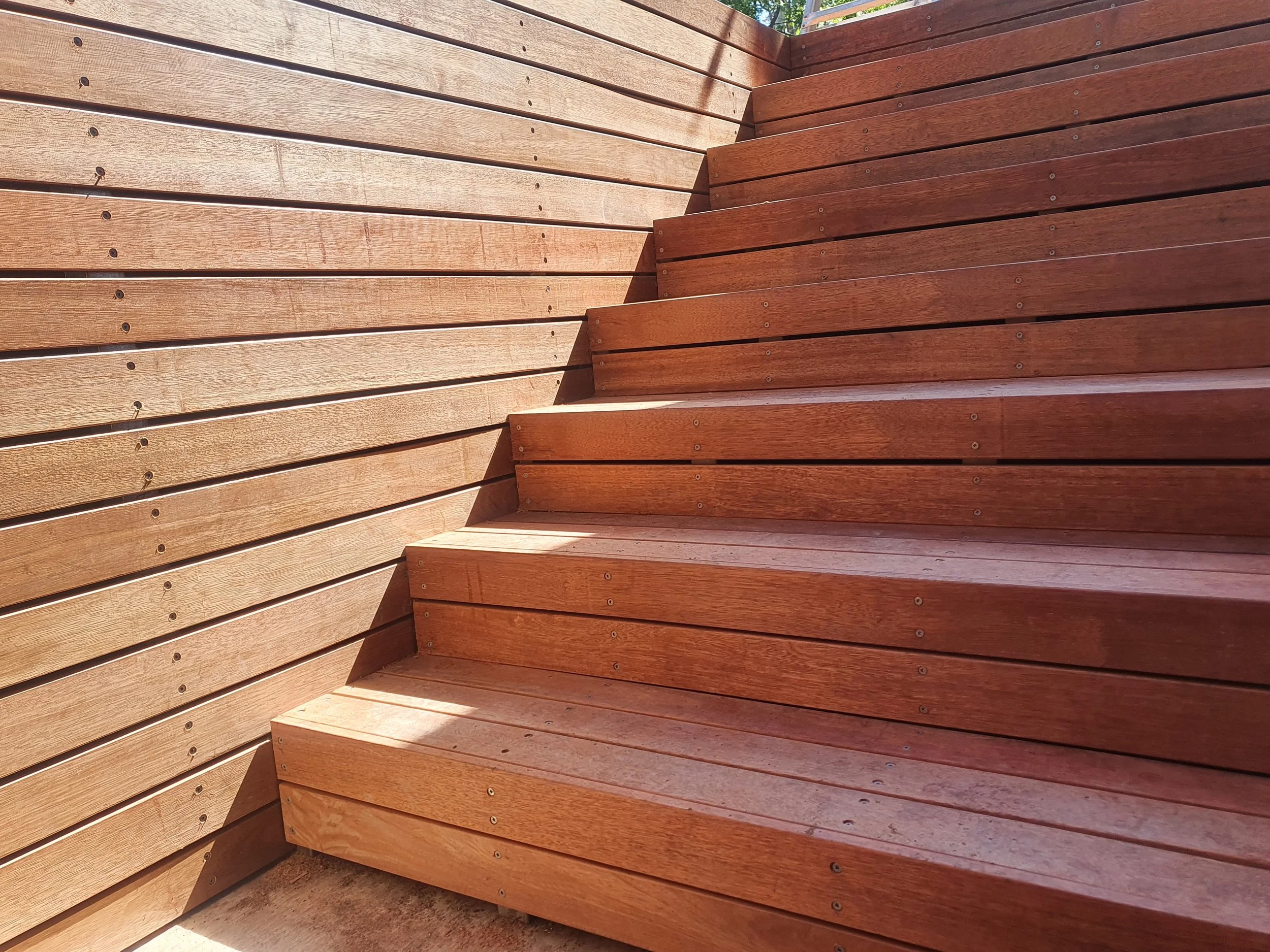 Close-up of a wooden staircase with natural and stained wood, showing parallel wooden planks with visible screws, taken outdoors during daytime.