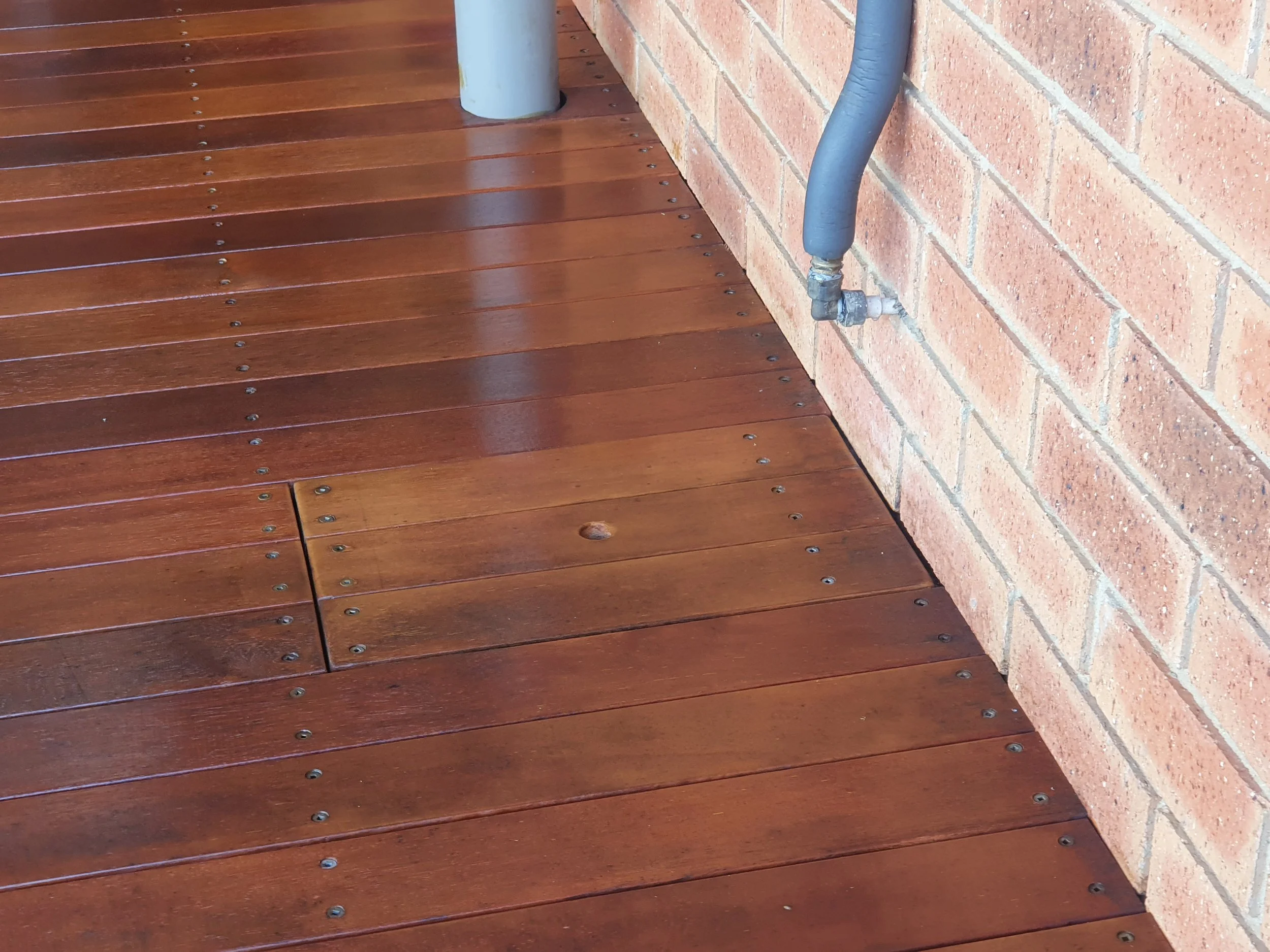 Close-up of wooden deck flooring with reddish-brown planks secured with screws, next to a brick wall with an outdoor hose attached to a water spigot.