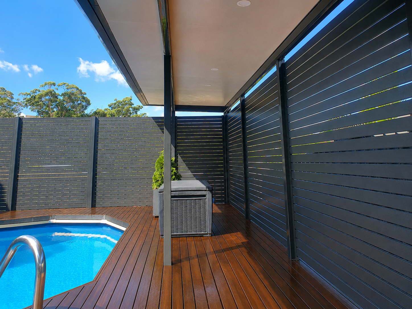 Luxury backyard with a small swimming pool, wooden decking, black privacy fence, and a modern outdoor seating area under a shaded overhang.