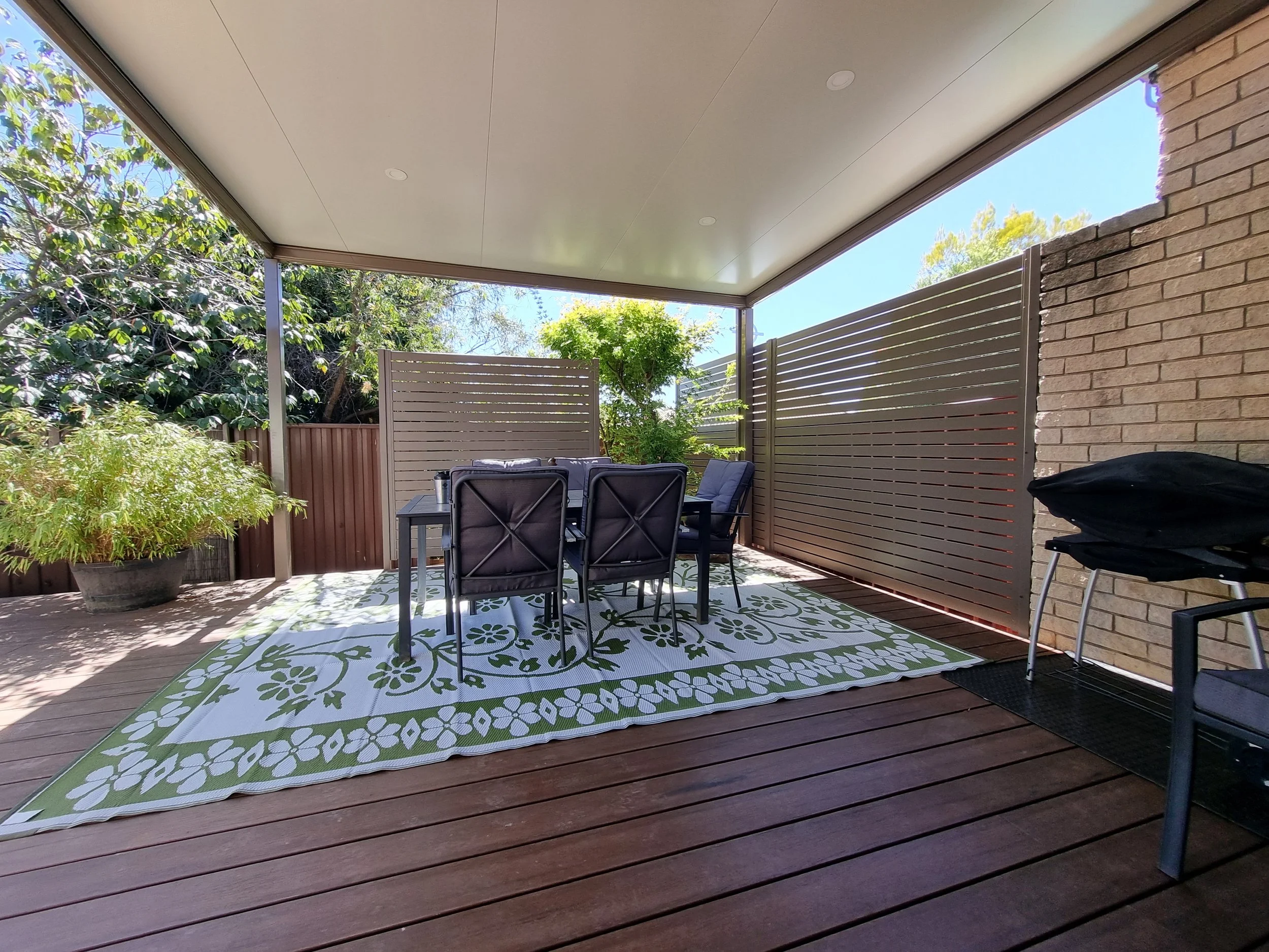 An outdoor patio area with wooden decking, a dining table with six chairs, a patterned outdoor rug, and greenery including trees and bushes behind a privacy fence, under a covered ceiling with recessed lighting.