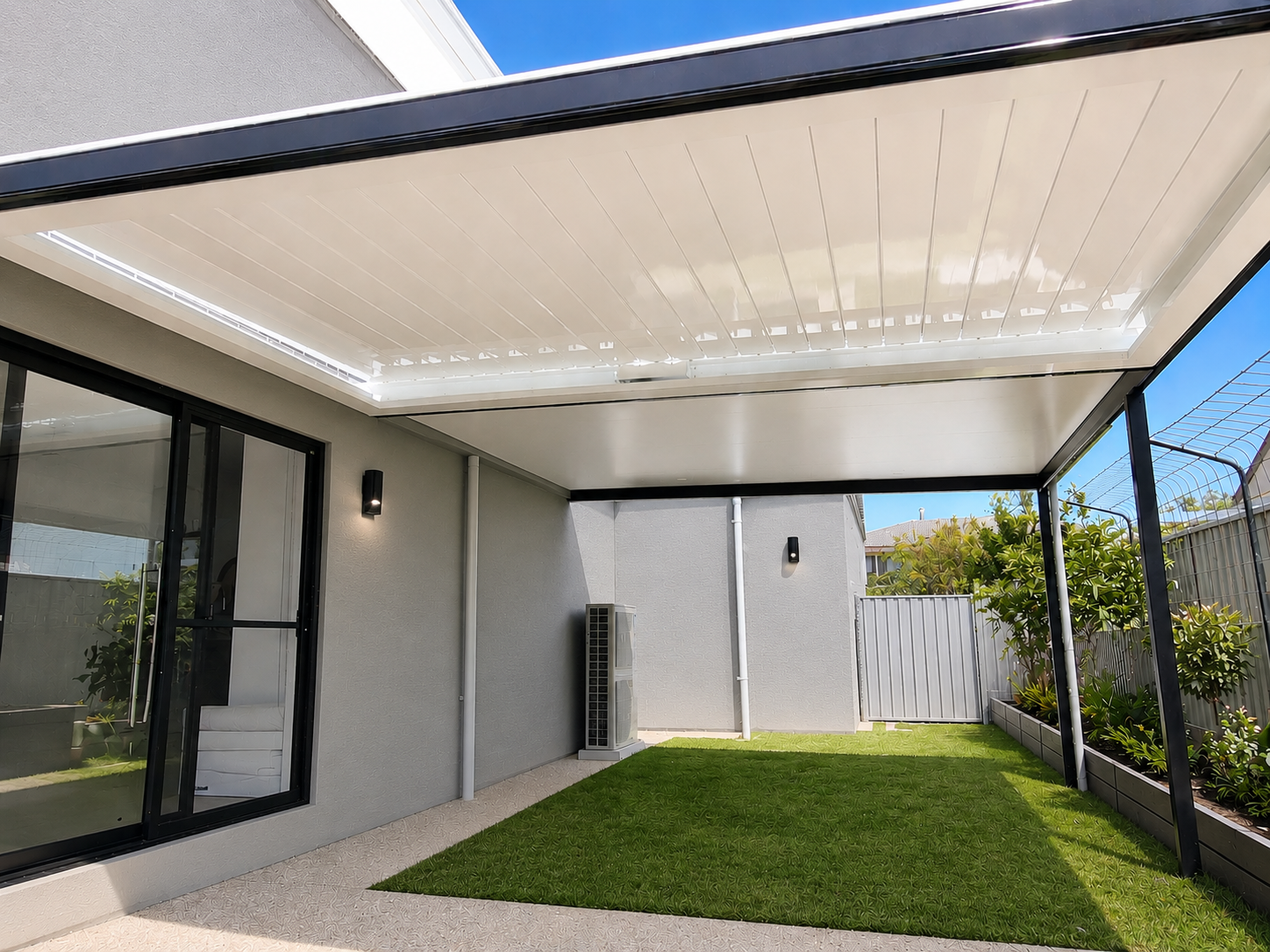 Covered backyard patio with artificial grass, sliding glass door, black outdoor wall lights, an air conditioning unit, and a garden bed with green plants, enclosed by a fence.