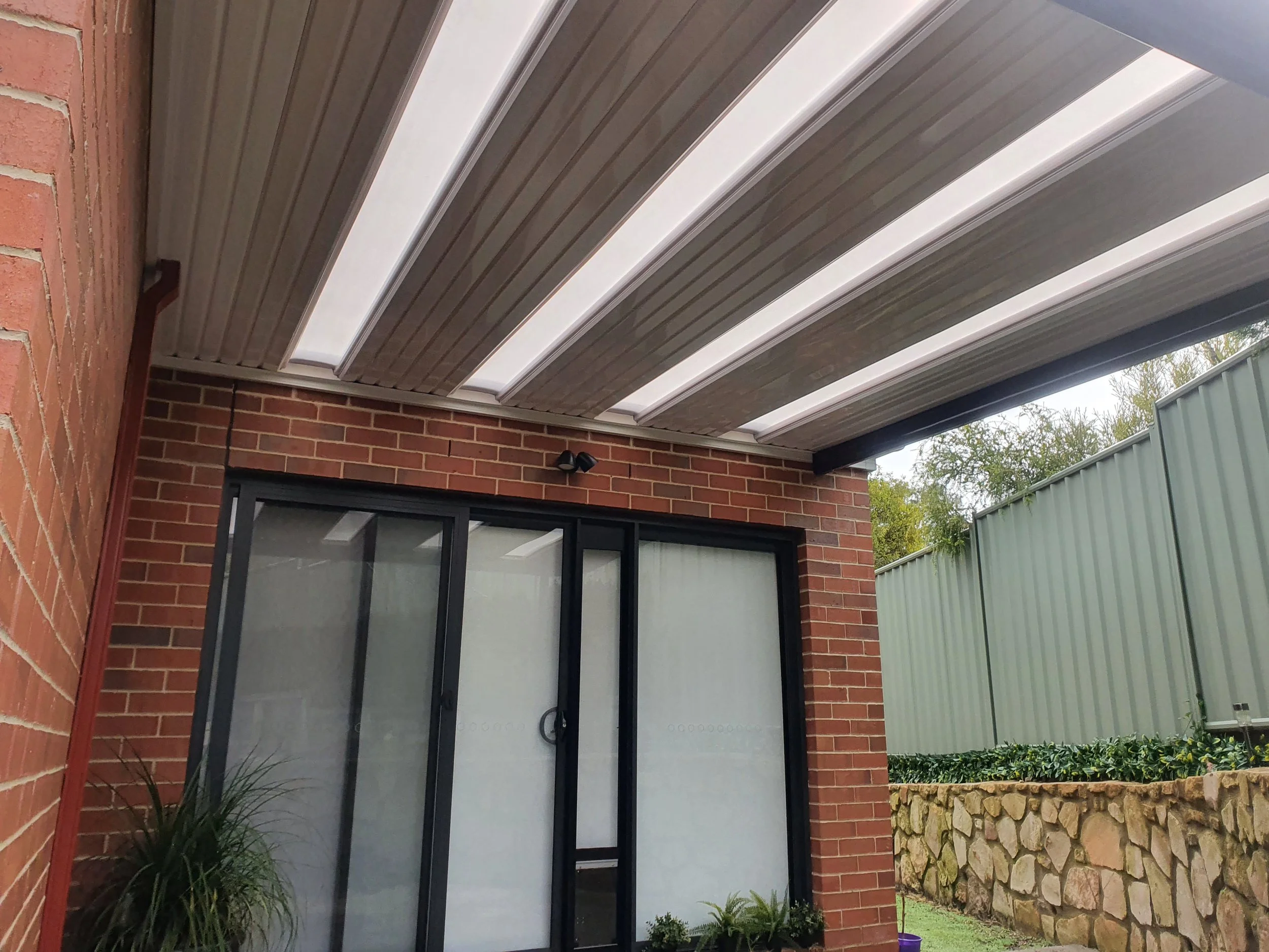 An outdoor residential area featuring a brick house with large sliding glass doors, a metal roof extension with translucent panels, and a garden with plants and a stone-bordered flower bed.