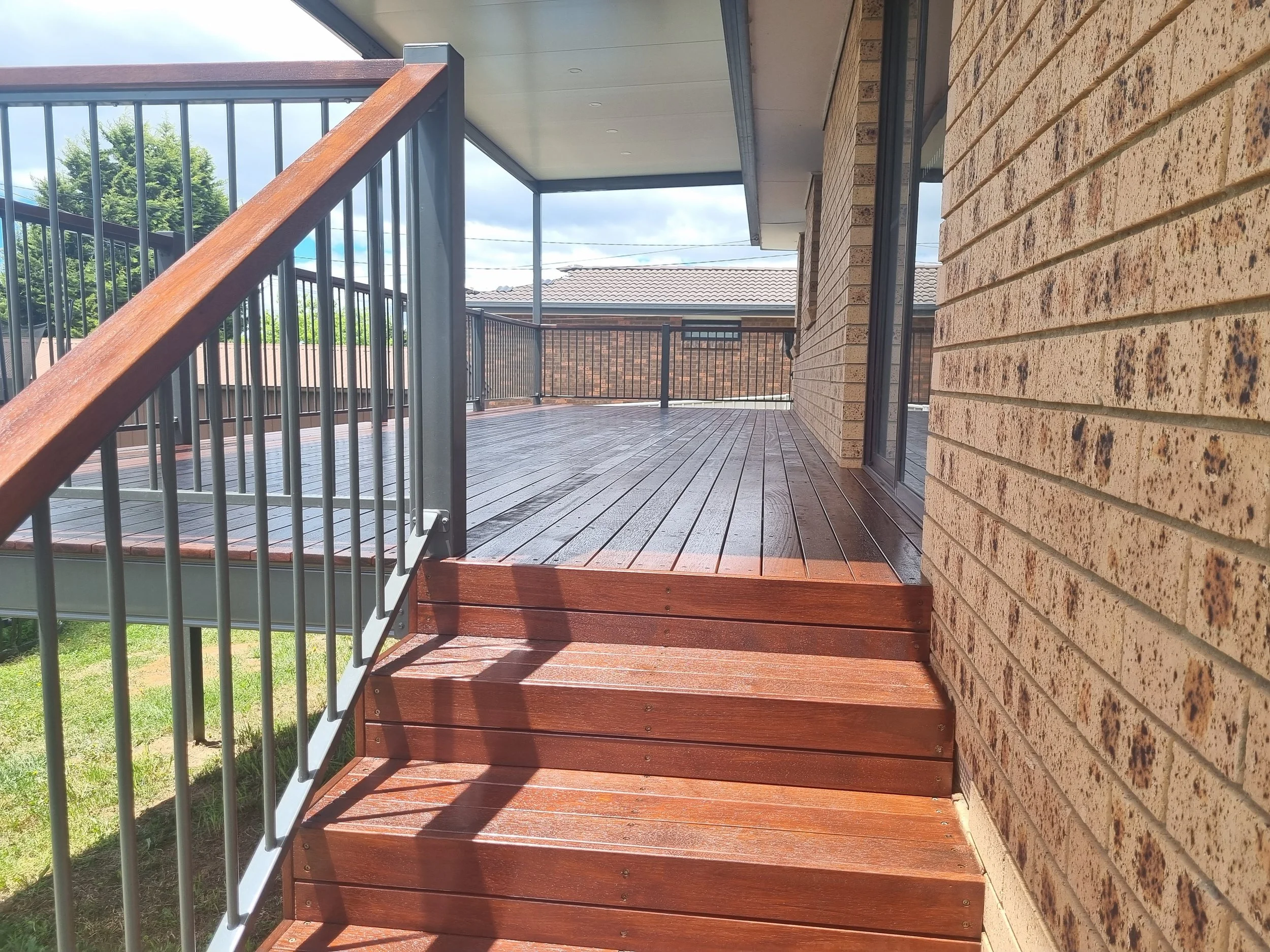 A wooden deck with stairs, metal railing, and brick wall on the right side. There's a house with a tiled roof visible in the background, and trees and sky beyond the railing.