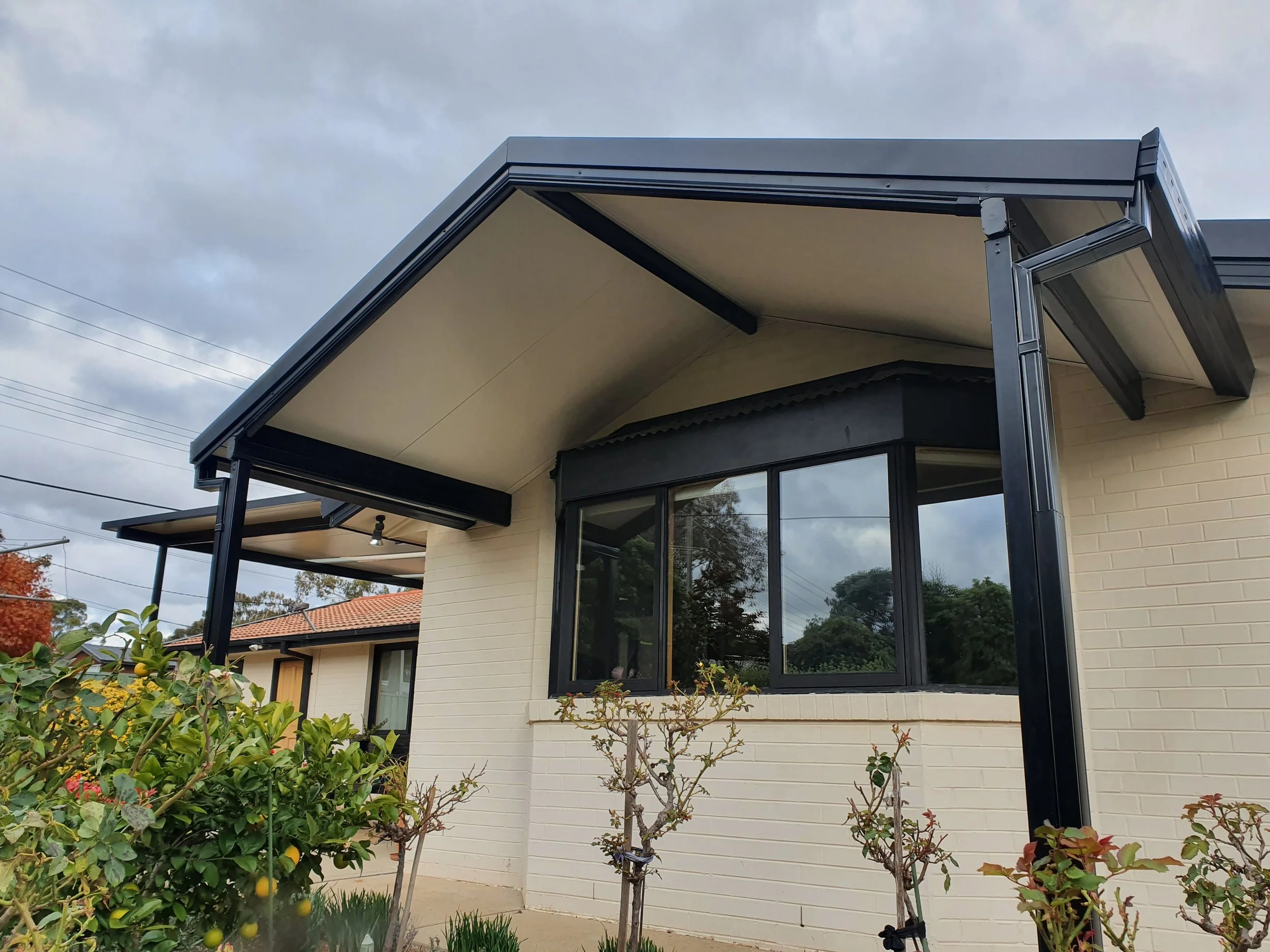 Modern house with black metal framing, large windows, and a sloped roof, surrounded by a garden with flowering plants and small trees under a cloudy sky.