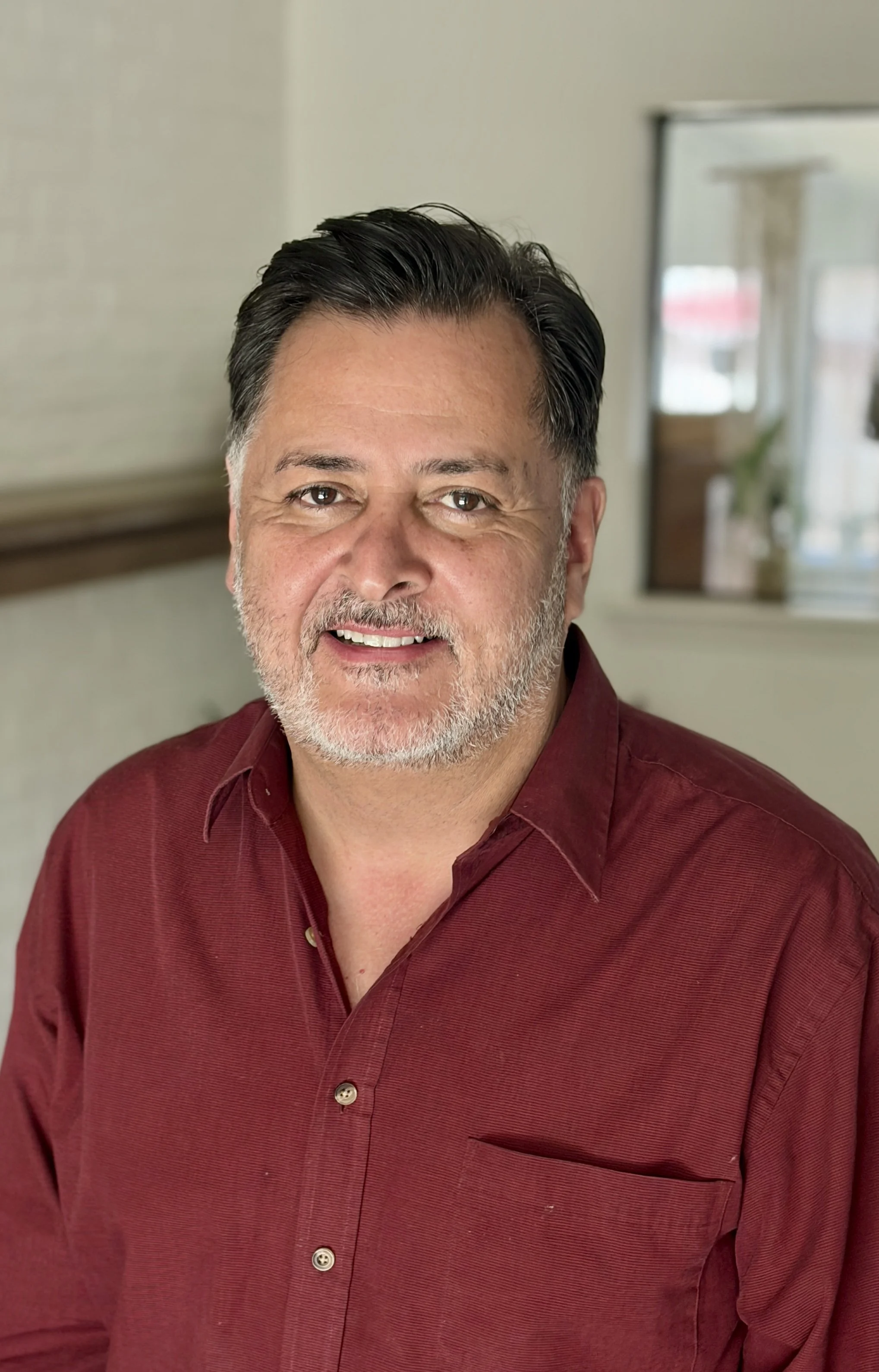 A middle-aged man with dark hair and a beard, wearing a red button-up shirt, smiling at the camera in a room with a window and a mirror in the background.