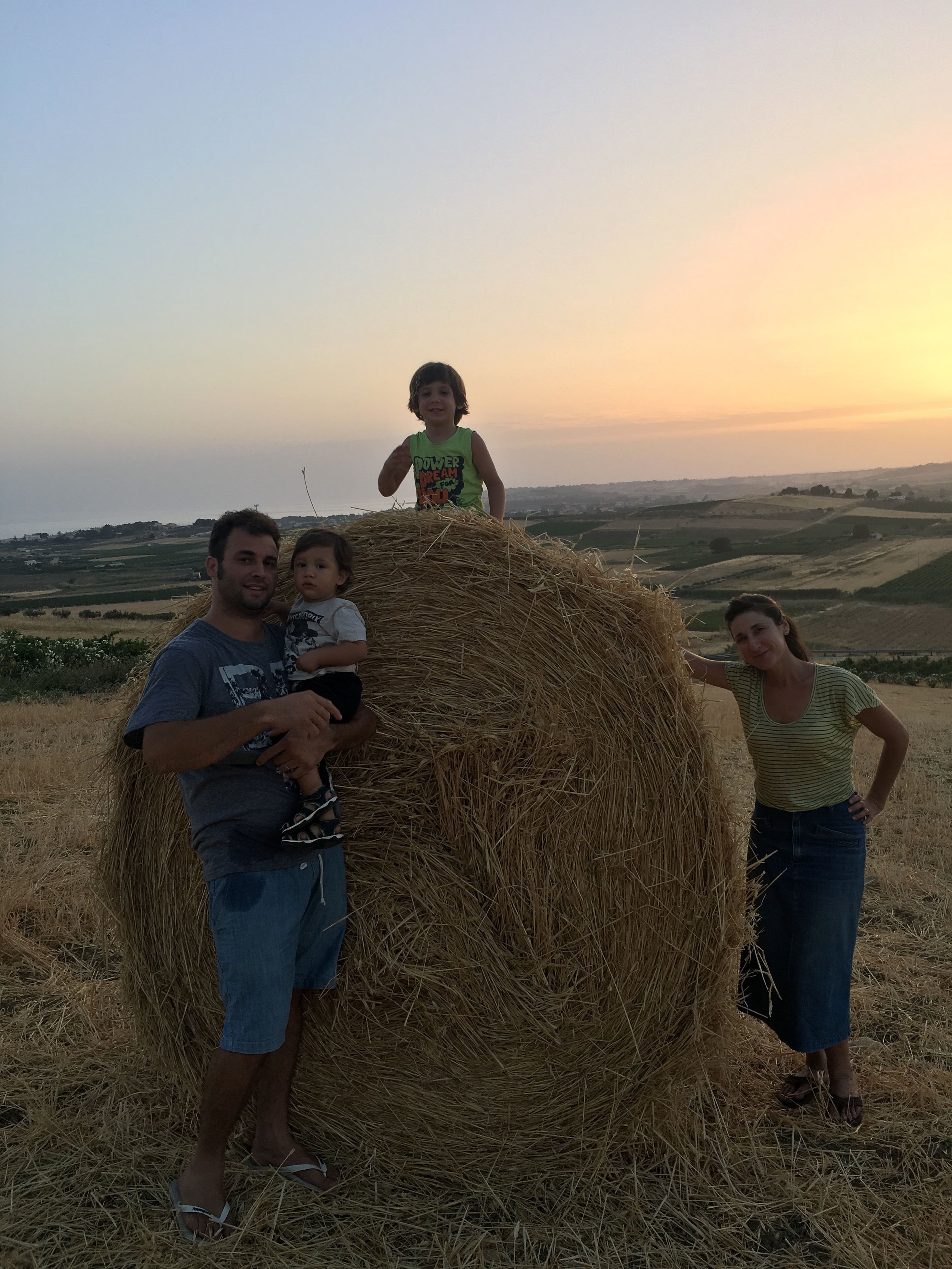 A family of four posing around a large hay bale in a rural landscape during sunset. agostino olive oil EVOO Menfi Sicily family 