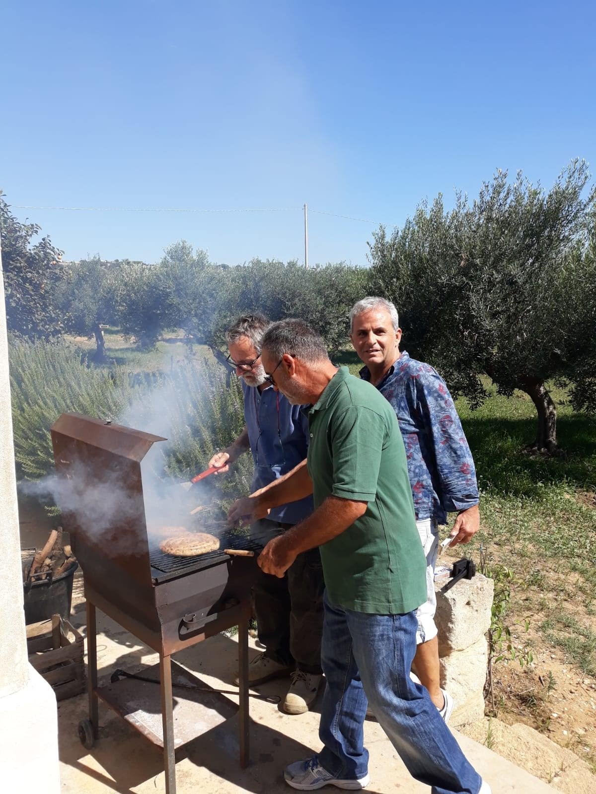 Three men are grilling hamburgers outdoors on a sunny day, with trees and a clear blue sky in the background. Lota's Family Agostino Olive oil EVOO