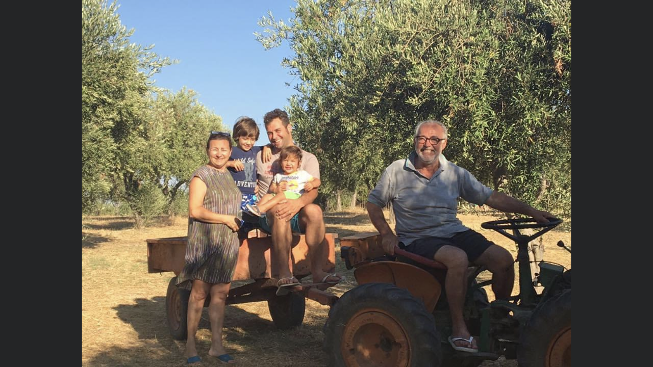 Lota's Family Grovee Olive. They are gathered around a person riding a tractor in a field with trees in the background. One woman stands next to the tractor, with the other family members sitting or standing nearby, smiling.