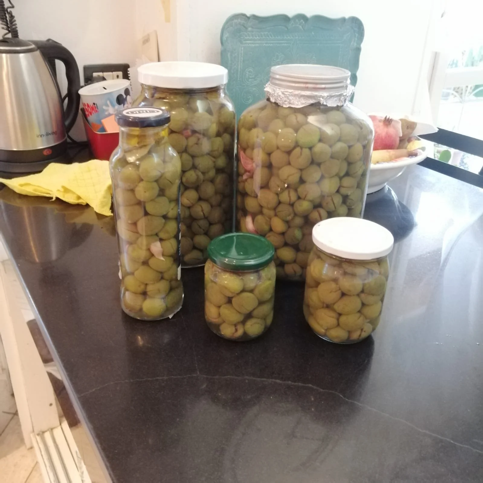 Several jars filled with greenish-yellow olives cured, likely pickled, . A yellow dish towel is also on the counter, along with a kitchen appliance and some other items in the background. EVOO cured olive 