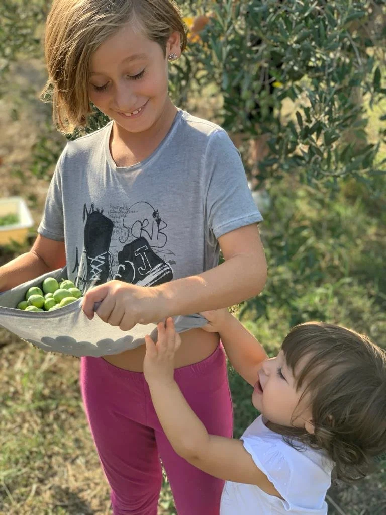 Two young girls outdoors, one older girl holding a bowl of green grapes and a younger girl reaching for the grapes with a happy expression.