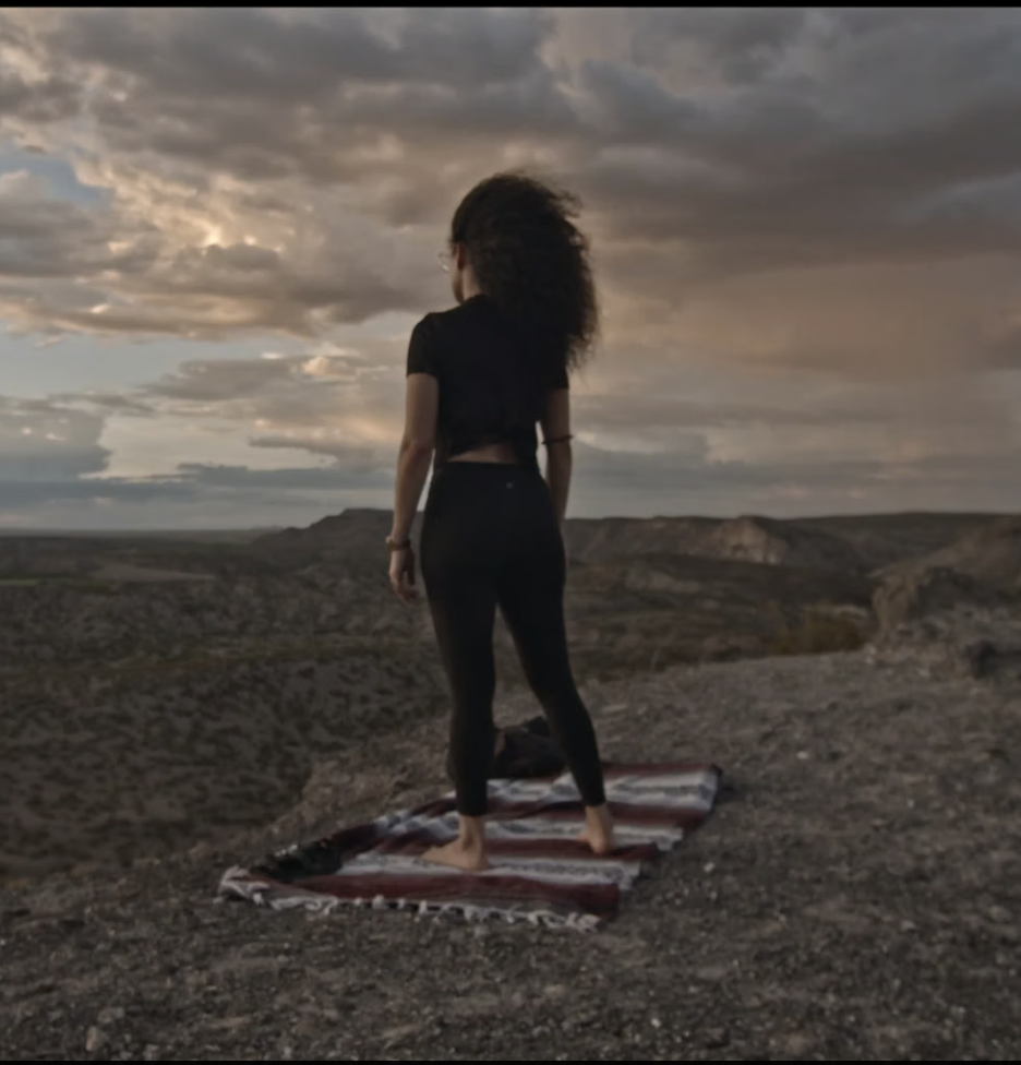 A woman standing on a patterned cloth outdoors during sunset, looking at a cloudy sky over a barren landscape.
