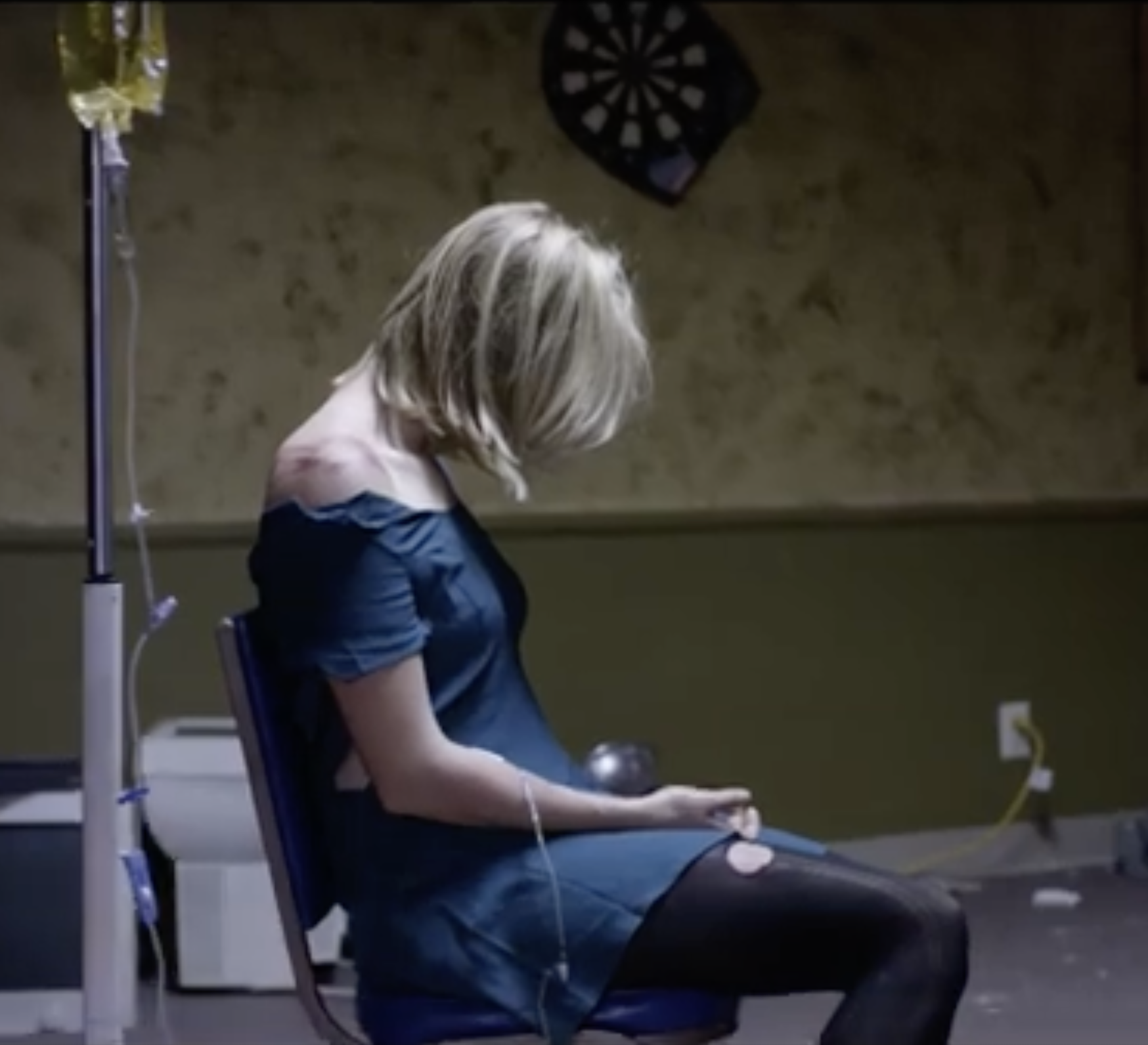 A woman with blonde hair sitting in a chair, in a medical or hospital setting, with IV equipment and wires nearby.