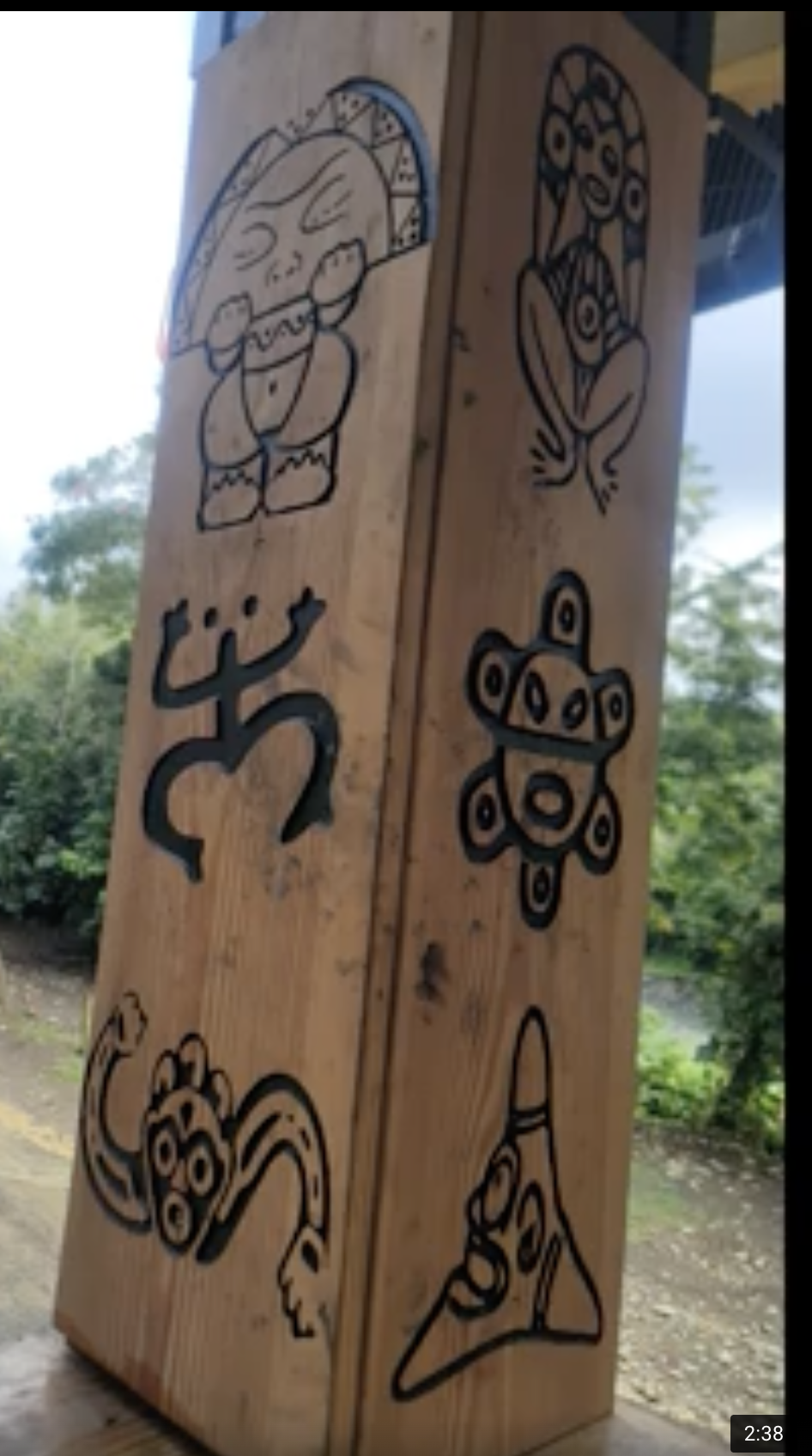 Wooden post with black line art tribal-style drawings of various animals and symbols, including a T-Rex, a lion, a turtle, a snake, and a hand pointing to a triangular symbol, with trees and sky in the background.