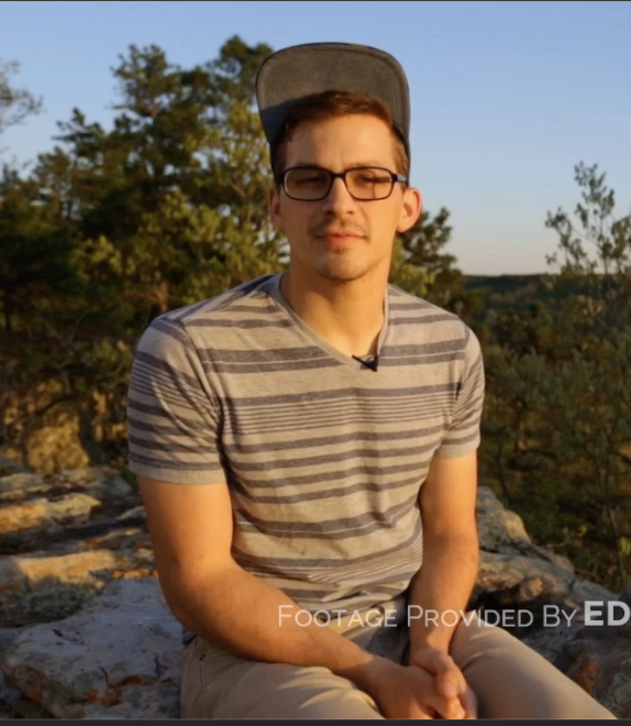 A young man wearing glasses, a striped T-shirt, and a baseball cap sitting outdoors on rocks during sunset with trees in the background.