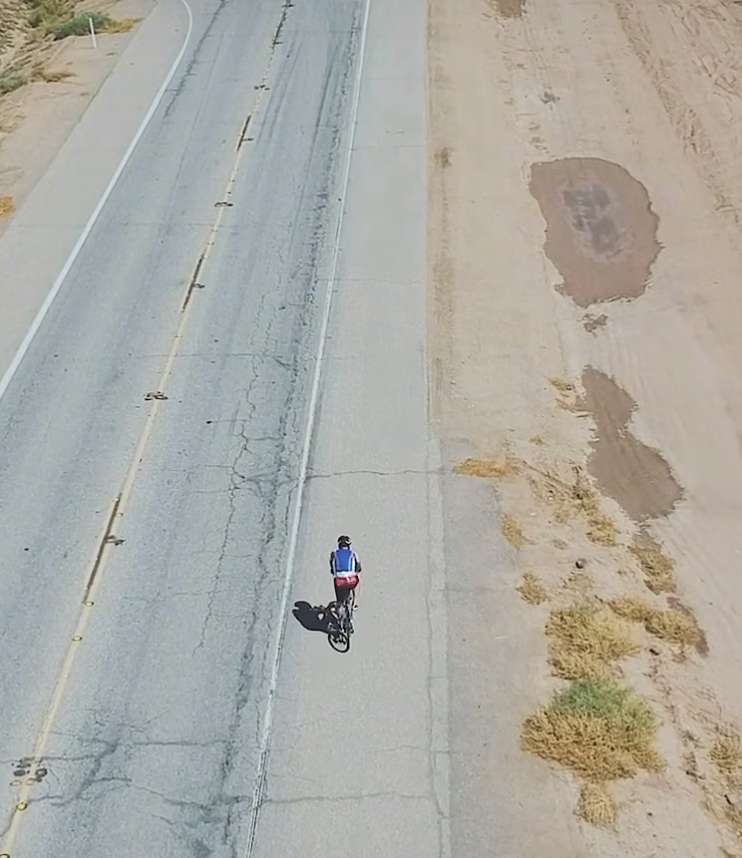 A person riding a bicycle on the sidewalk beside a cracked, empty road with patches of dirt and sparse vegetation.