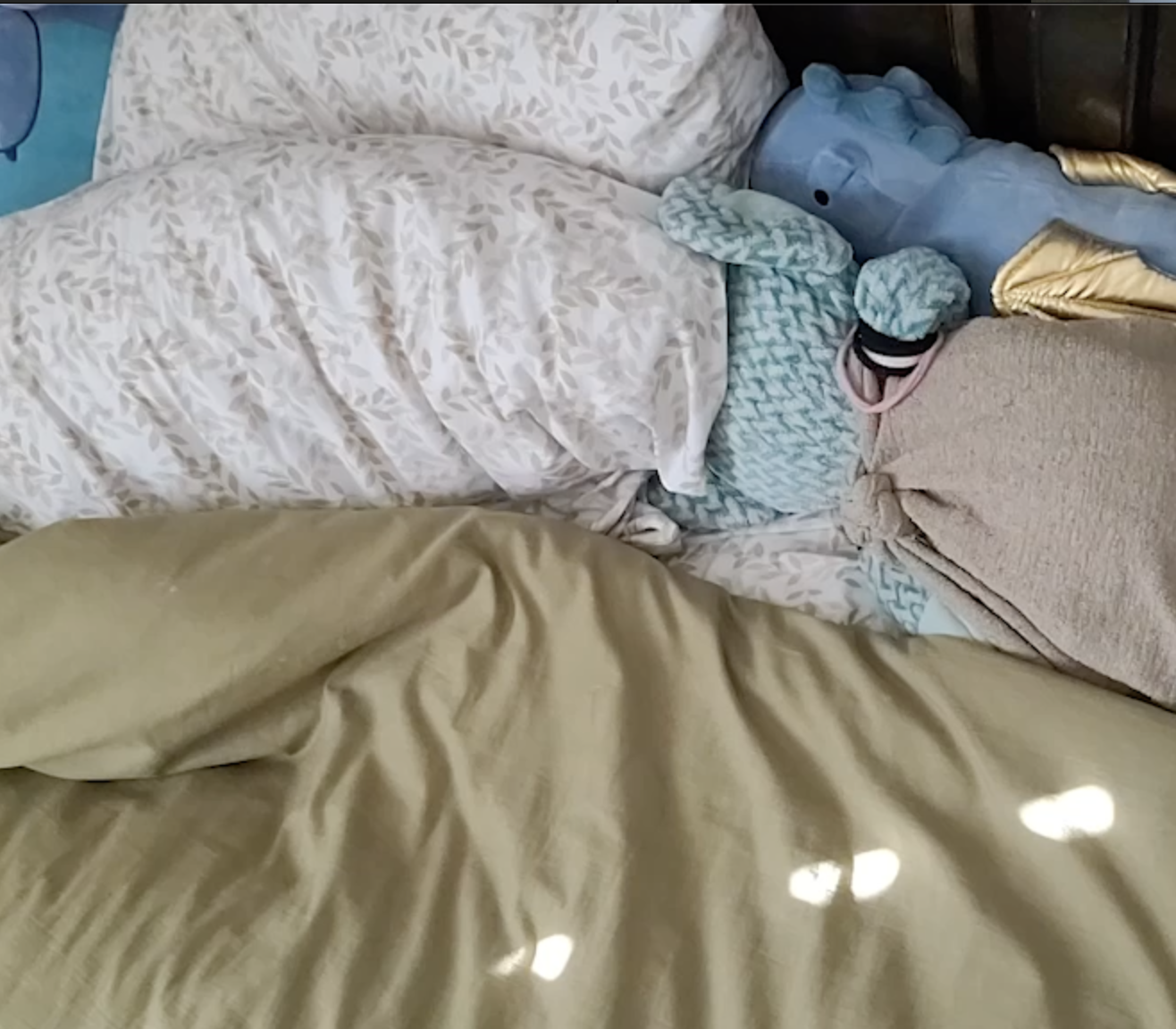 Disorganized bed with various blankets and stuffed animals, including a blue giraffe and a teal patterned blanket, with sunlight reflections on the beige bedspread.