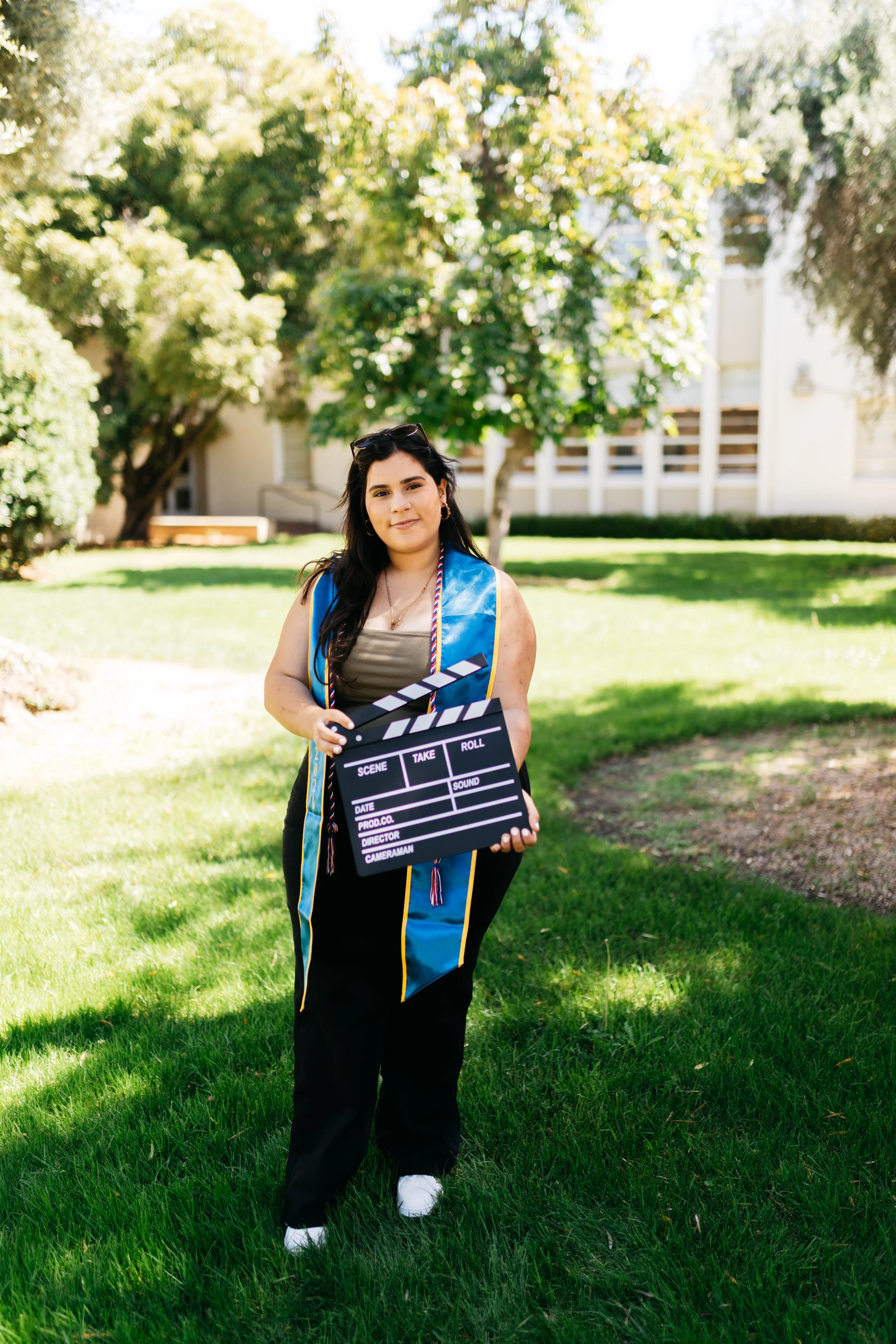 Young woman in graduation gown holding a clapperboard outdoors on a sunny day with trees and a building in the background.