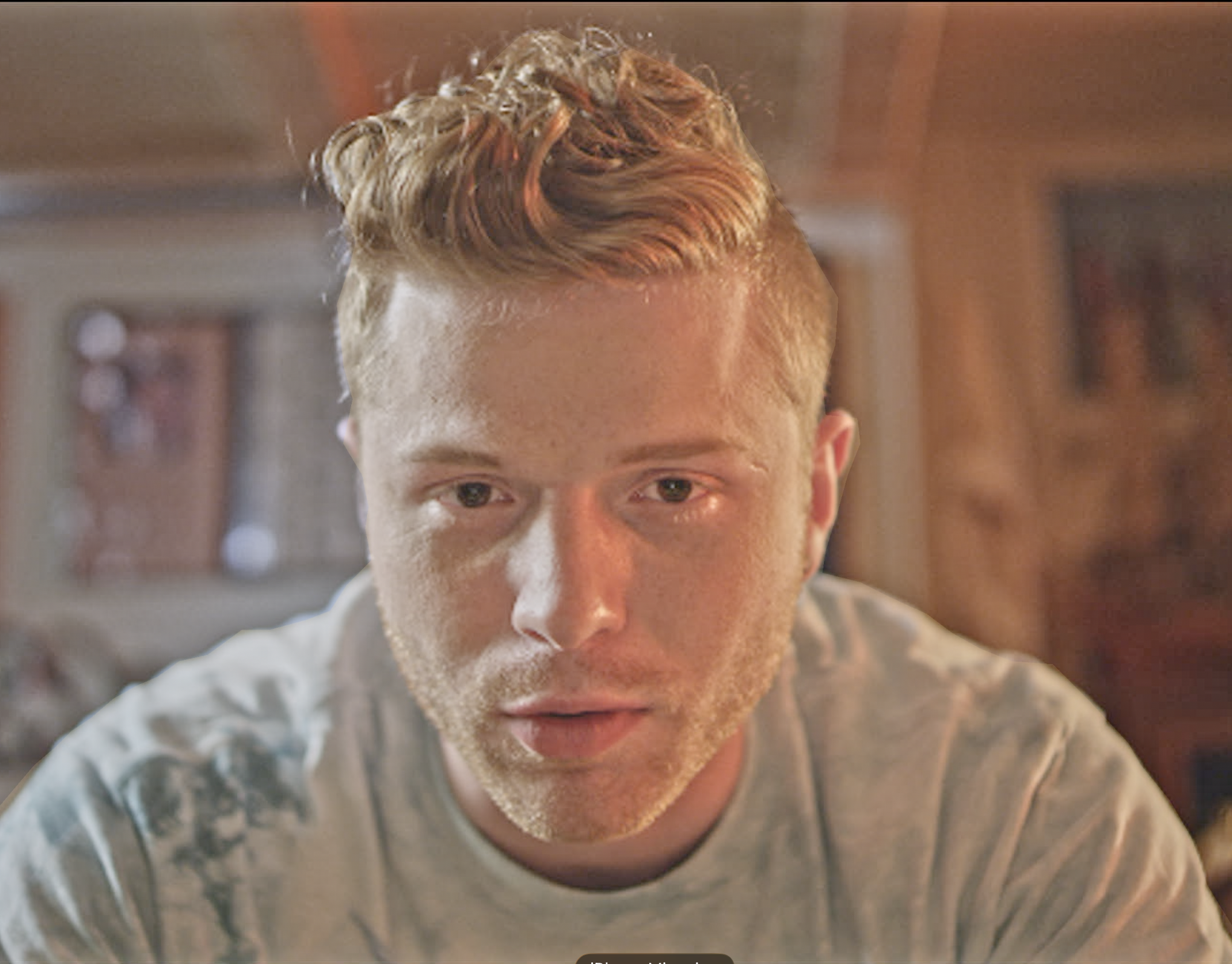 A young man with light skin and curly blonde hair, indoor setting, wearing a light gray shirt, looking down slightly, with soft lighting.