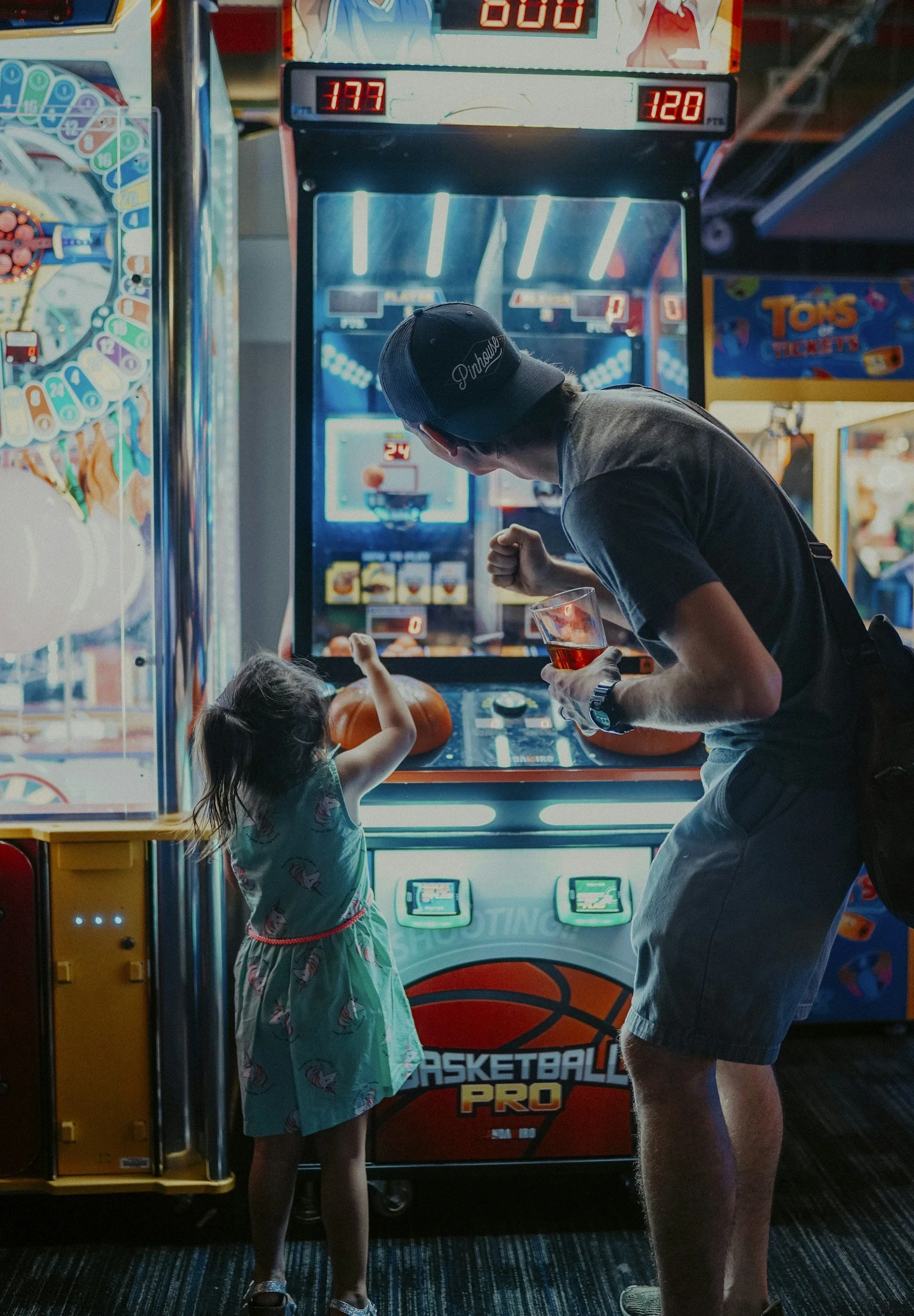 A man and a young girl playing an arcade basketball game in an arcade filled with various game machines and bright lights. The girl points towards the game while the man leans in holding a drink.