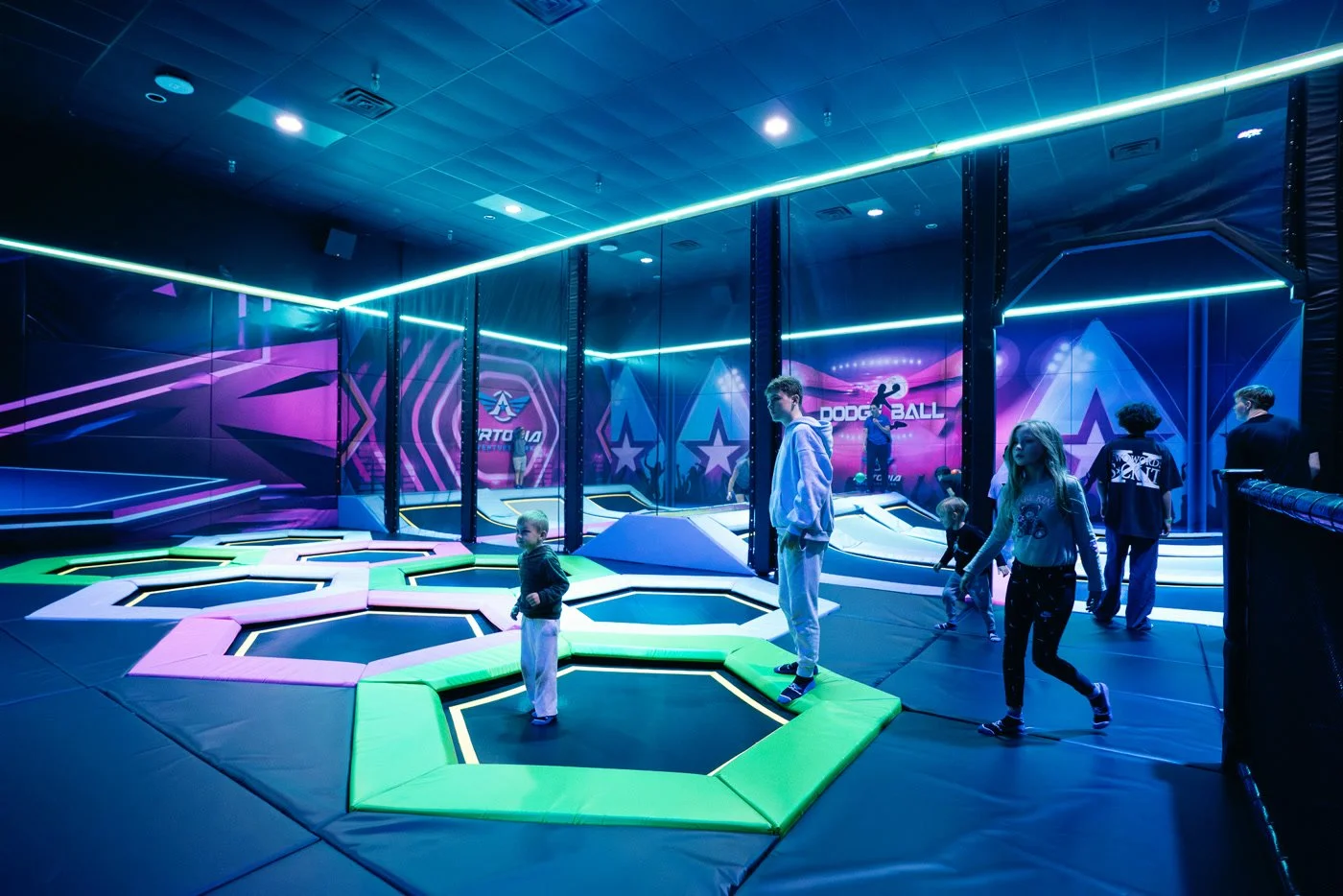 Indoor trampoline park with children jumping on colorful trampolines, neon lighting, and a Dodgeball area with neon signs.