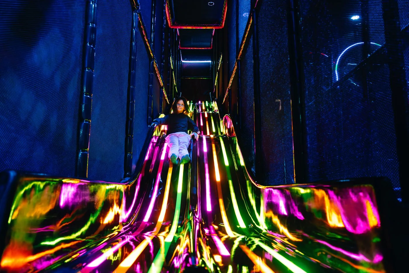 Child riding down a colorful, neon-lit slide in an indoor playground.