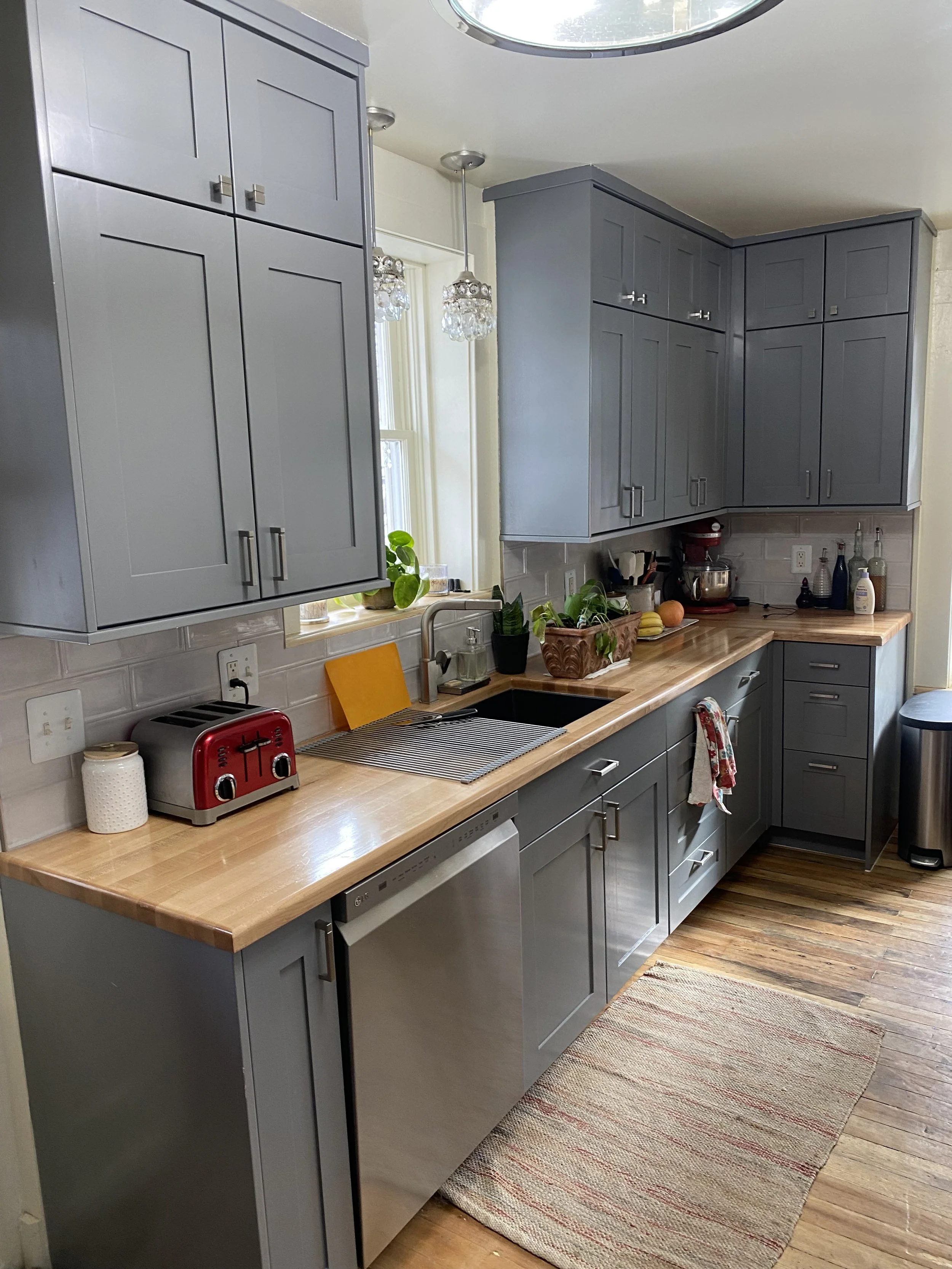 A kitchen with gray cabinets, a wooden countertop, a red toaster, potted plants, a window with hanging chandeliers, and various kitchen items on the counter and shelves.