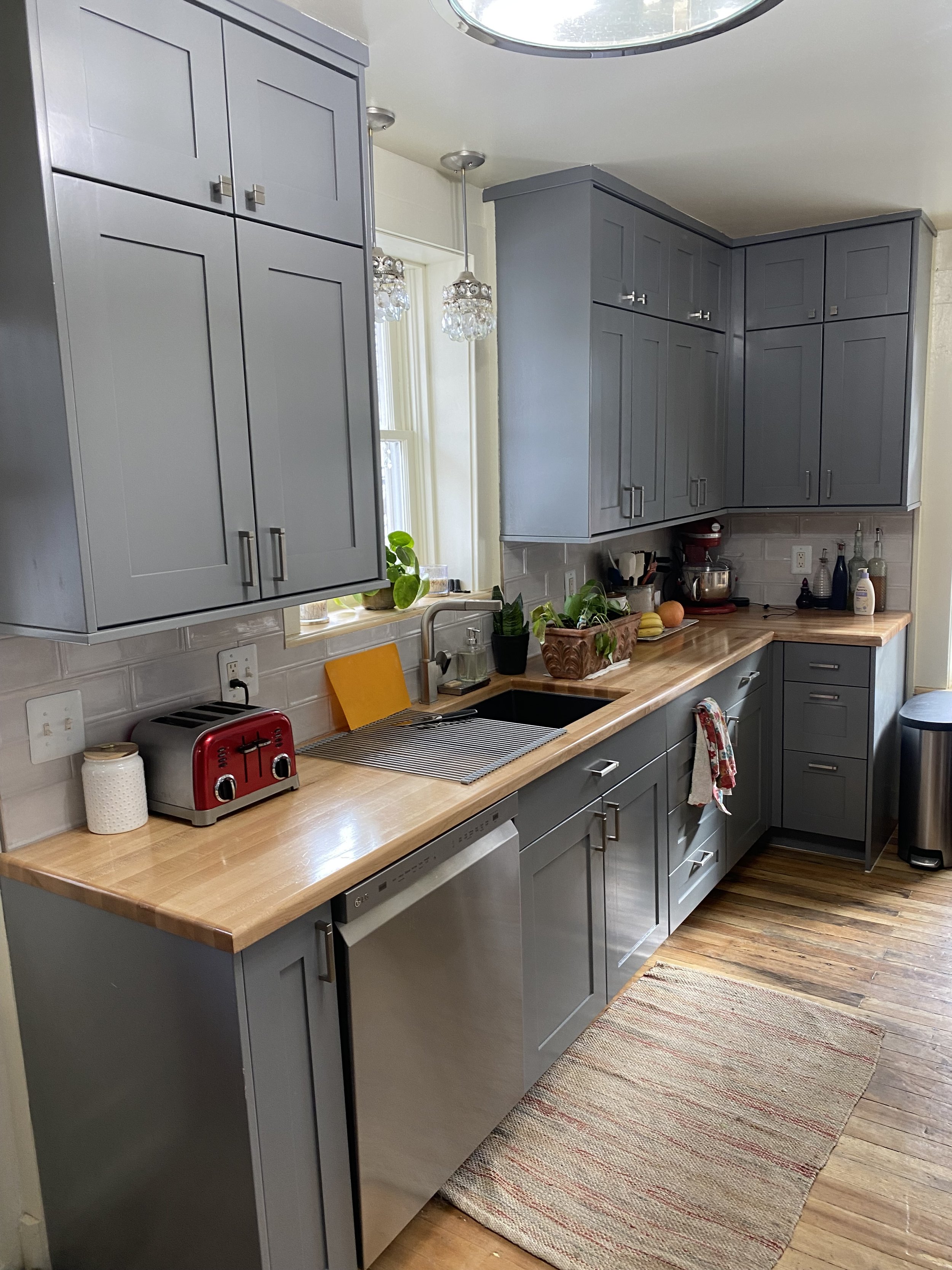 A kitchen with gray cabinets, a beige wooden countertop, a window with plants, a red toaster, and various kitchen items.