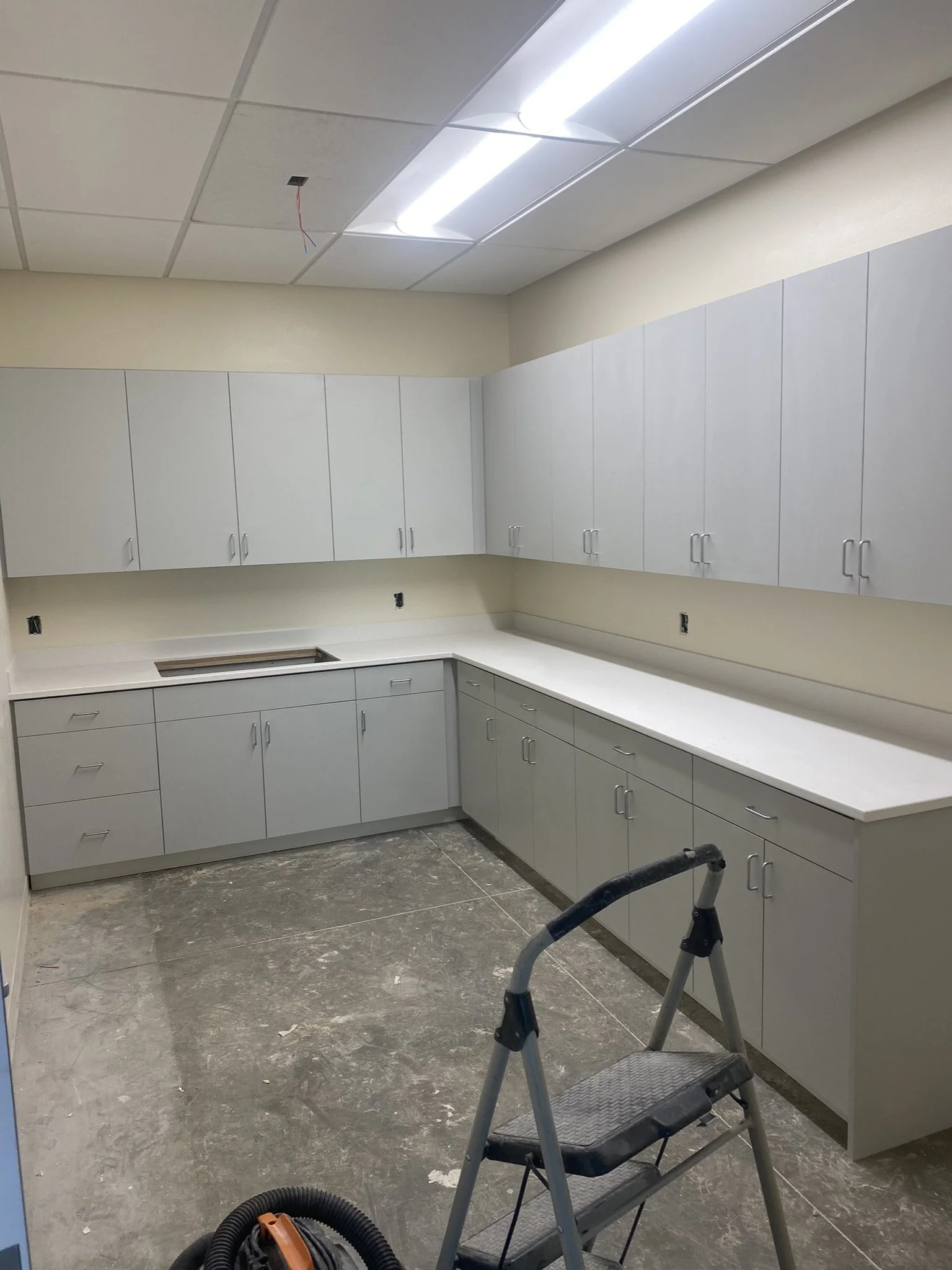 Construction or renovation scene in a room with white cabinets and a partially installed ceiling. There is a ladder, a vacuum cleaner, and construction tools on the unfinished floor.