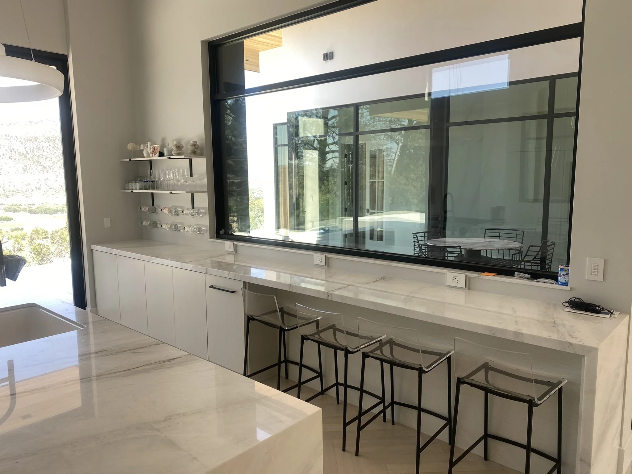Modern kitchen with white marble countertops, black bar stools, large windows showing a backyard with trees, and a shelving unit with cups and glassware.