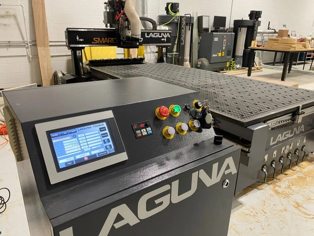 Industrial CNC machine labeled 'Laguna' with control panel, emergency stop button, and various knobs, in a manufacturing workshop.