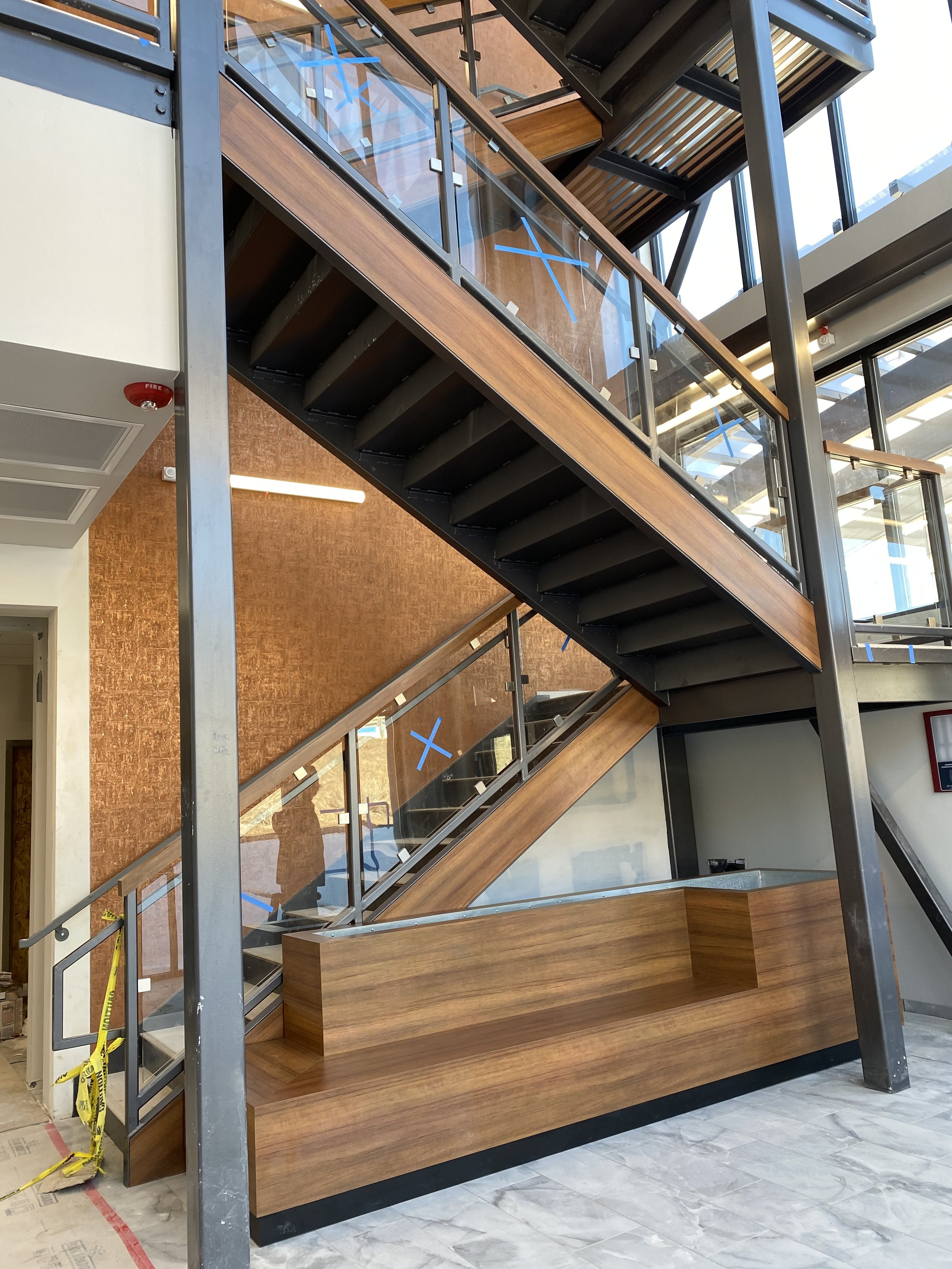 Modern interior staircase with glass railings, wooden steps, and metal framework in a building under construction or renovation.