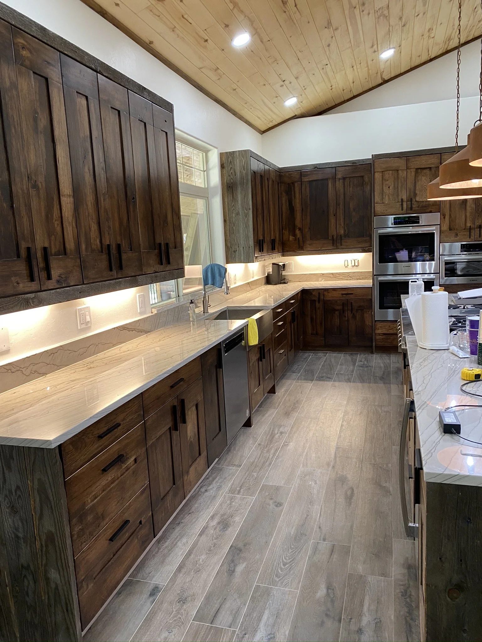 A modern kitchen with dark wooden cabinets, a marble countertop, stainless steel appliances, a window above the sink, and a wooden ceiling with recessed lighting.