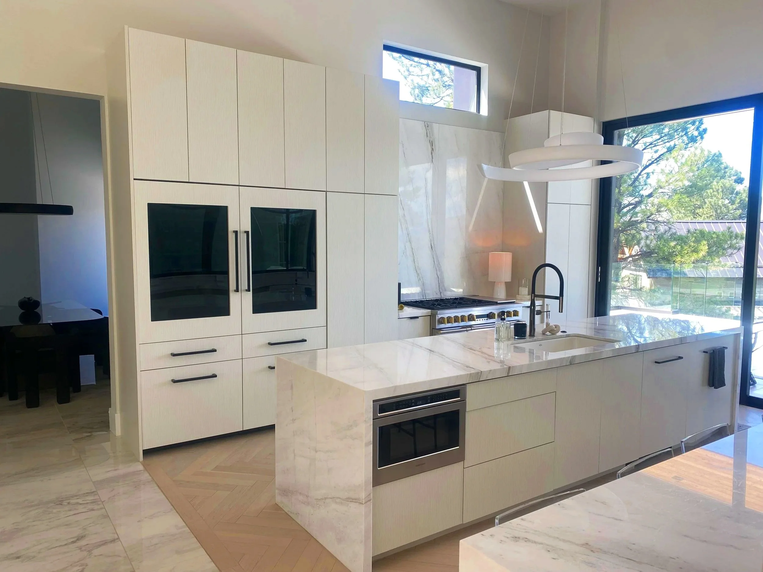 Modern kitchen with white cabinetry, marble countertops, large windows, and a circular white pendant light fixture.