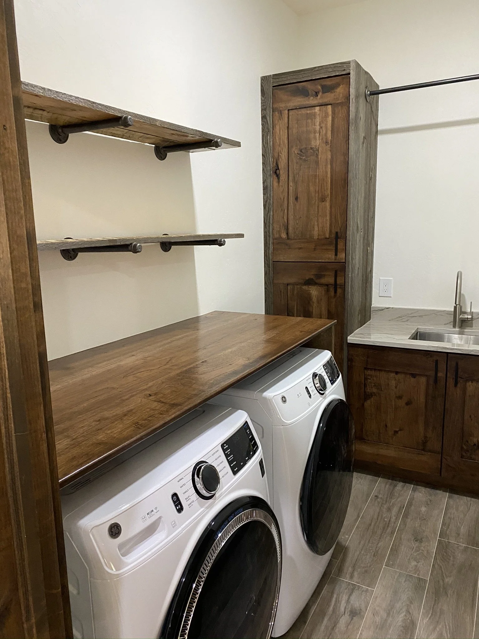 A laundry room with a washing machine and dryer side by side, wooden countertops above, open shelving, a wooden cabinet, and a sink.