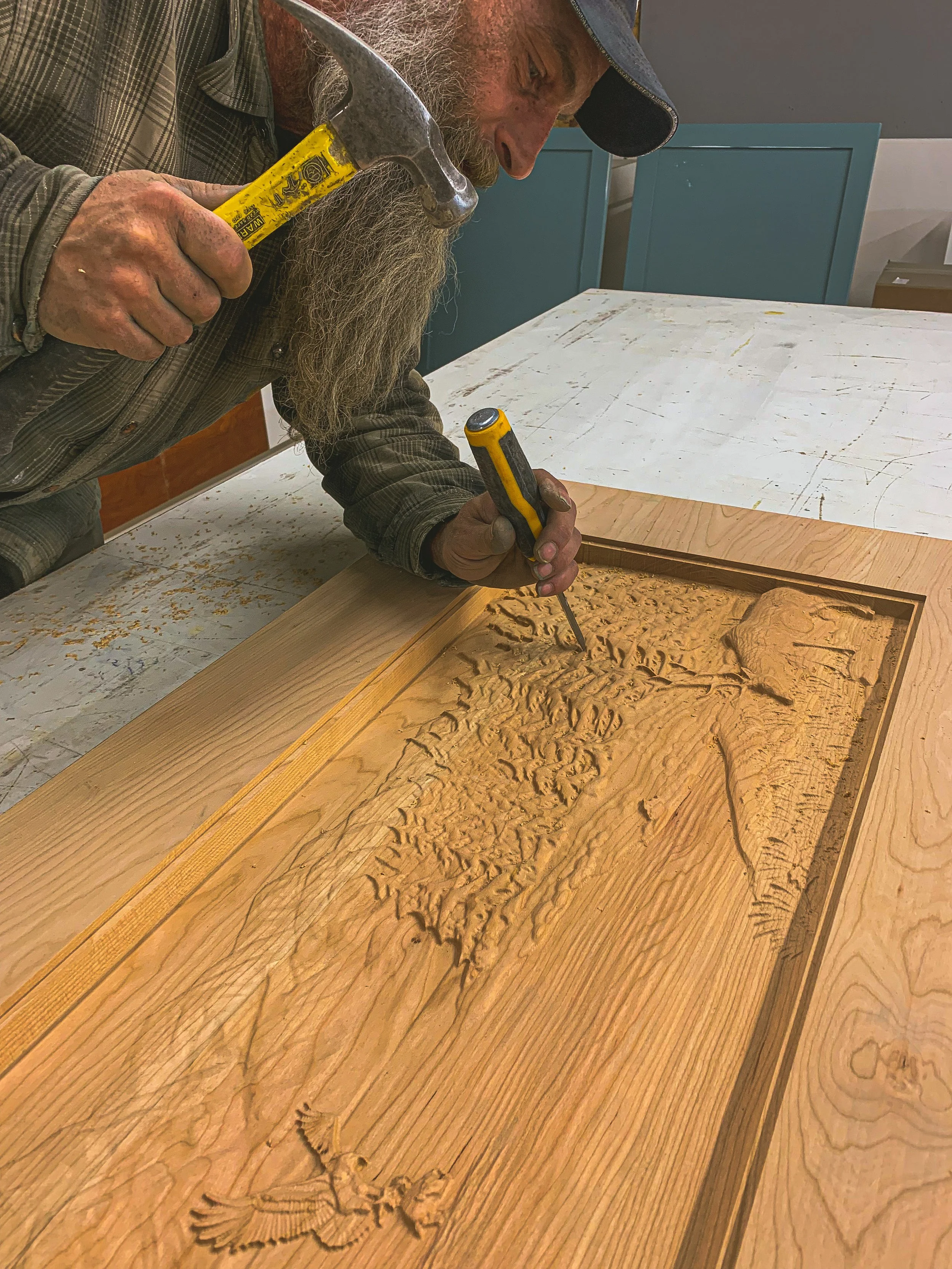 A man with a long beard and wearing a plaid shirt and a baseball cap is using a chisel and hammer to carve intricate patterns into a piece of wood on a workbench.